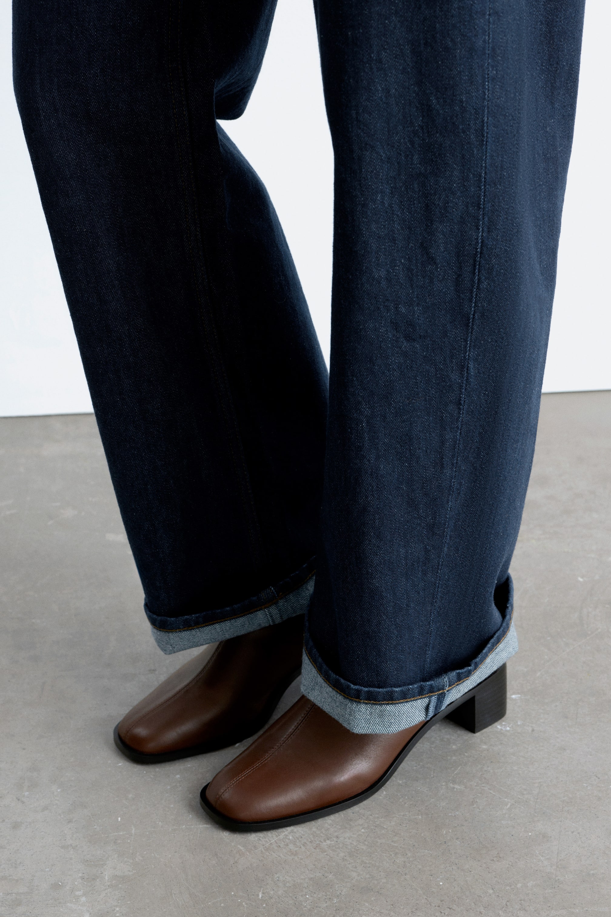 Grotere afbeelding bekijken: Donkere denim jeans zijn omgeslagen bij de enkels, waardoor een lichtere denim voering zichtbaar wordt. Ze worden gedragen over bruine leren enkellaarsjes met vierkante neuzen en donkere blokhakken.