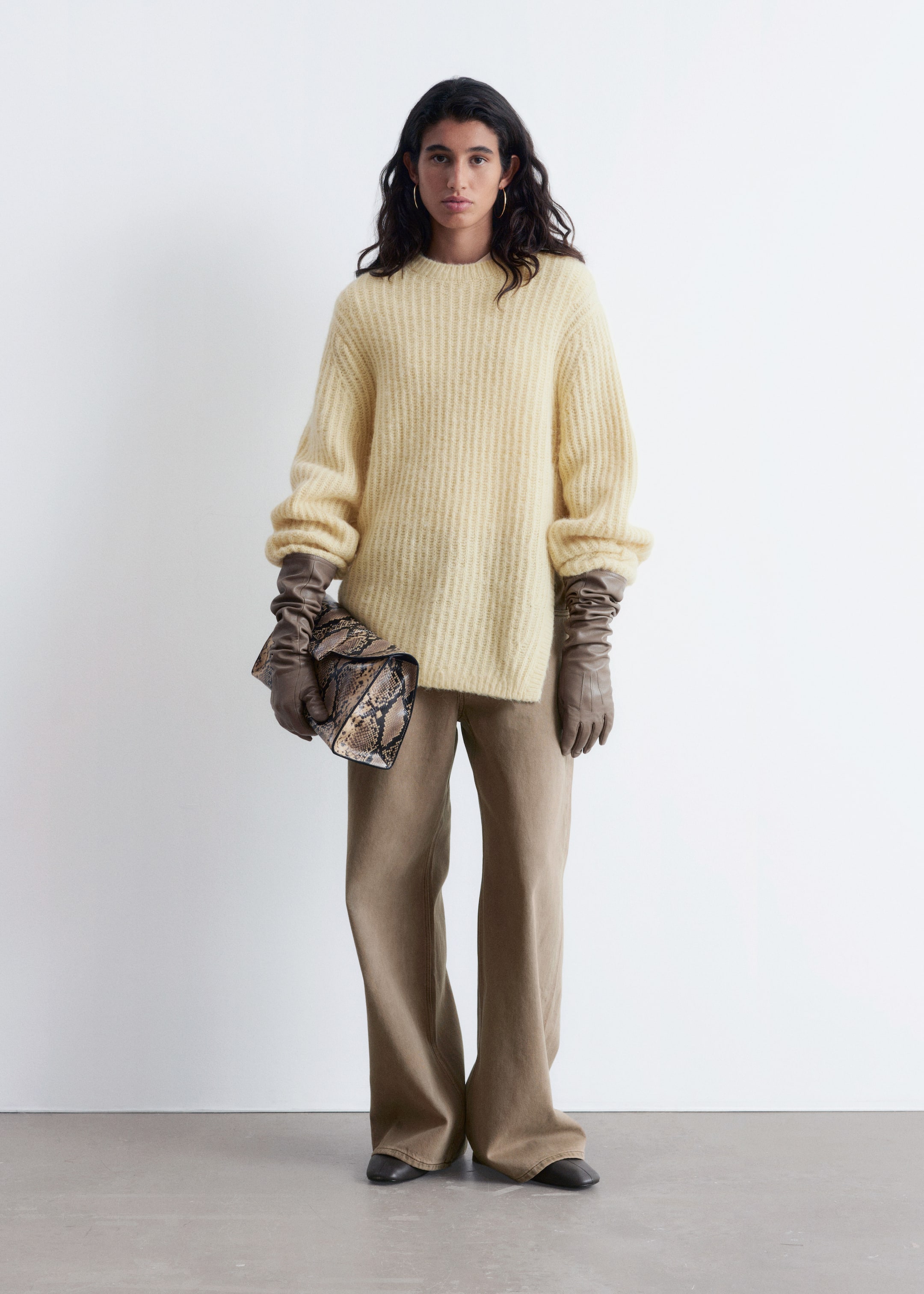 Visualizza immagine più grande: Un ensemble presenta un maglione a maglia a coste oversize di colore giallo chiaro con scollo a girocollo e maniche lunghe e voluminose che si raccolgono ai polsi. È abbinato a pantaloni color cachi a gamba larga e a tutta lunghezza e guanti lunghi in pelle marrone arricciata. Una borsa a mano con stampa pitonata viene tenuta in mano, e delle scarpe basse nere completano il look.
