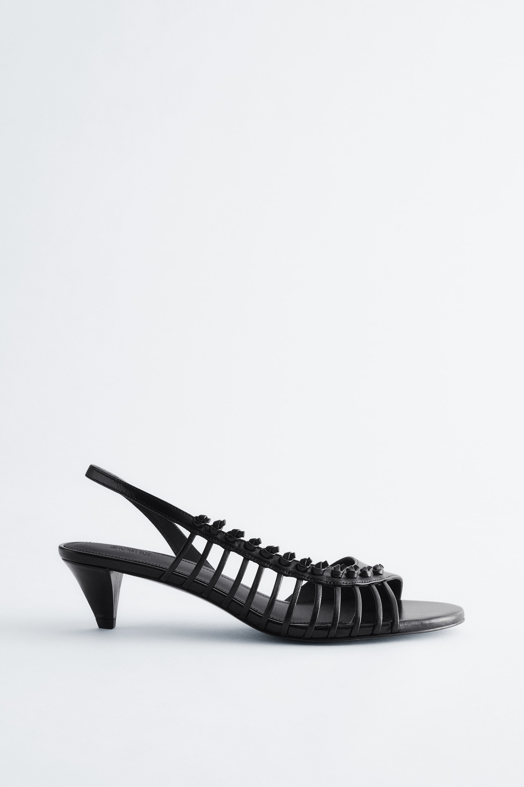 Größeres Bild anzeigen: Ein schwarzer Slingback-Sandal steht nach links, mit offener Zehenpartie und einem Käfigriemendesign über dem Rist, verziert mit kleinen, konischen Nieten. Ein schmaler Slingback-Riemen sichert die Ferse, die auf einem ausgestellten Kitten-Heel ruht.