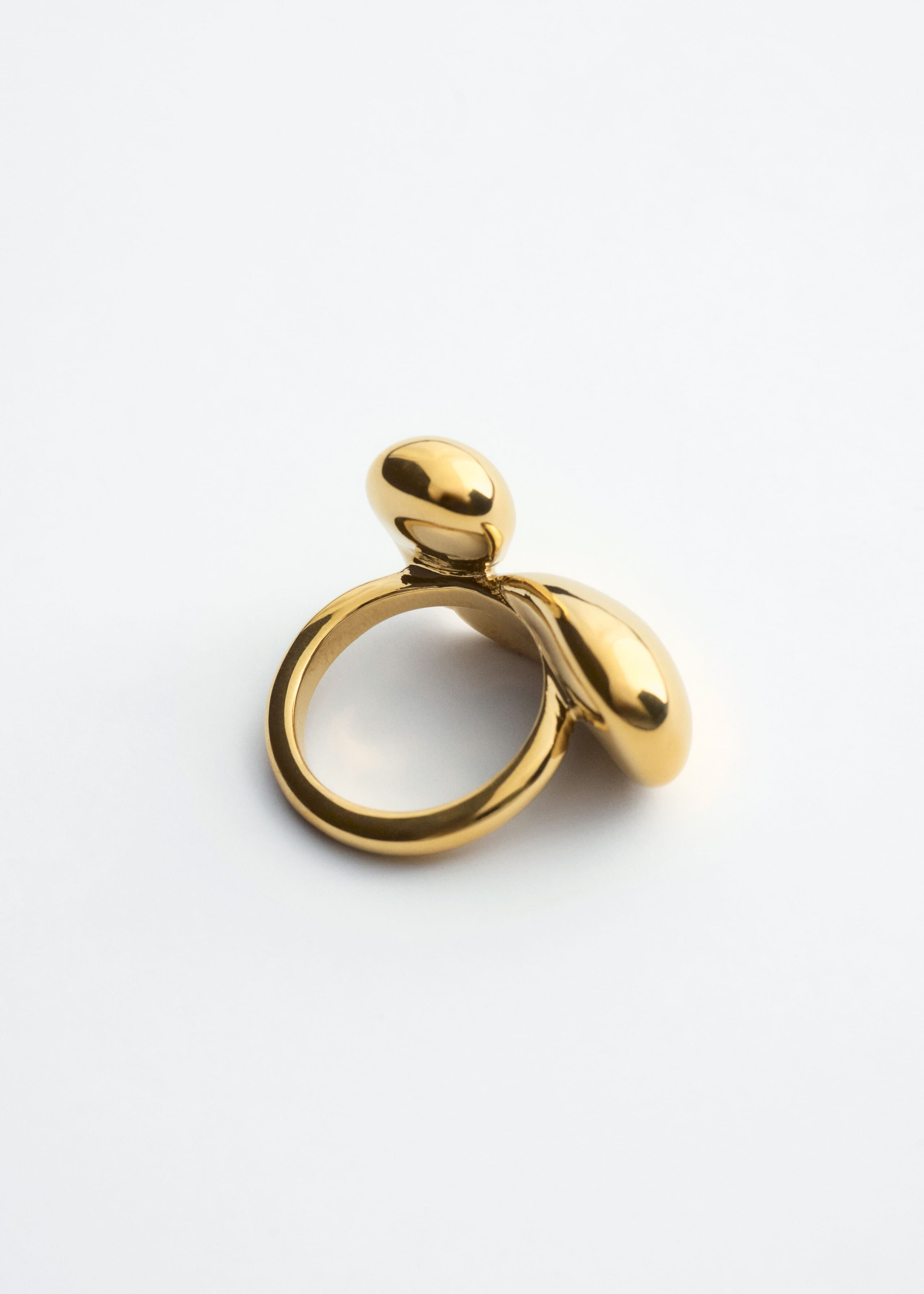 Un anello scultoreo in ottone dal tono dorato è appoggiato in diagonale, mostrando il retro del suo design con una fascia circolare liscia. Due forme fluide, bulbose e simili a ciottoli si estendono dalla parte superiore della fascia, mettendo in risalto una finitura altamente lucida e riflettente.