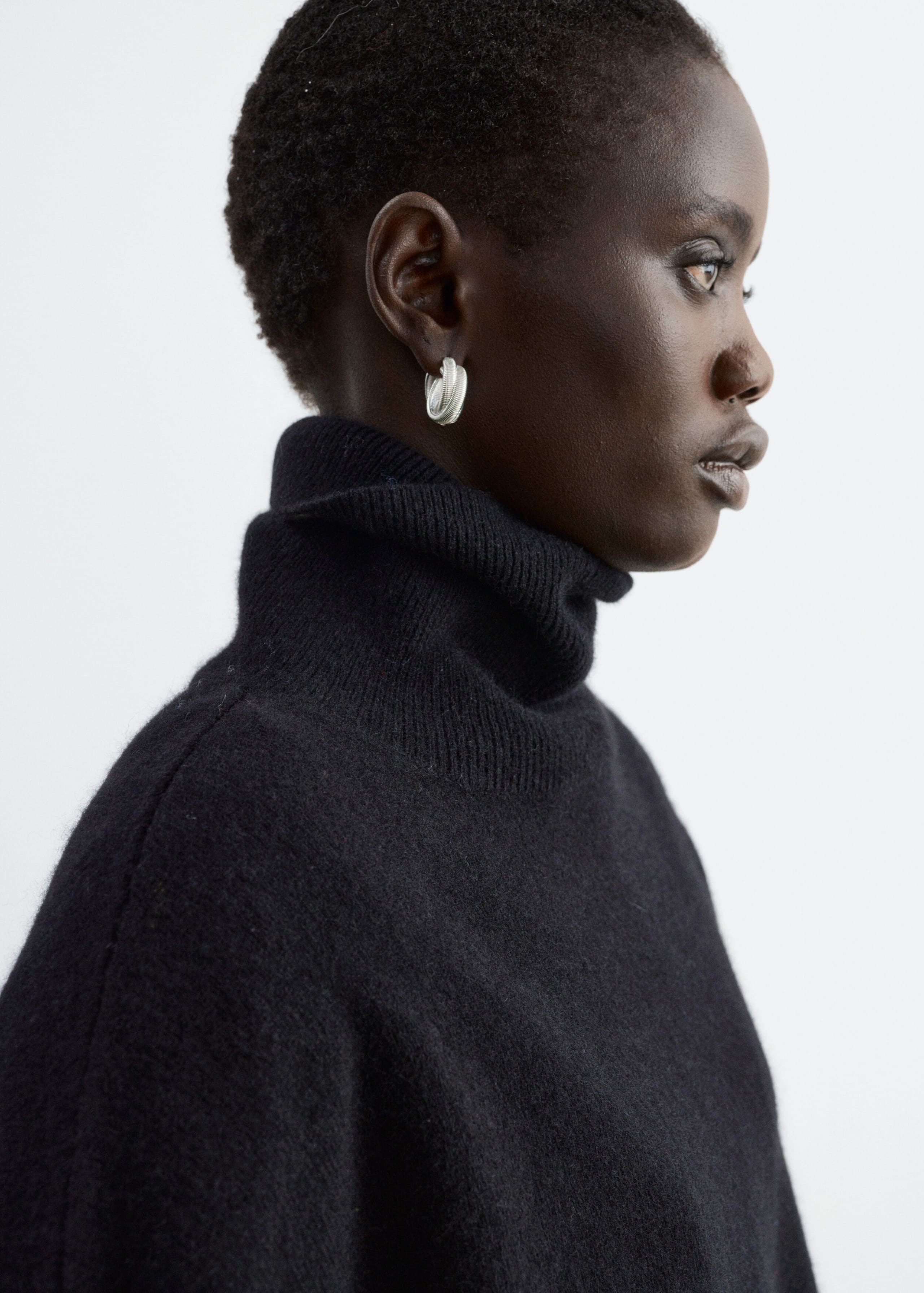 A black knit jumper featuring a folded ribbed roll neck and dropped shoulders is being worn by the model in a side-profile view. The soft, textured fabric is styled with a small silver hoop earring.