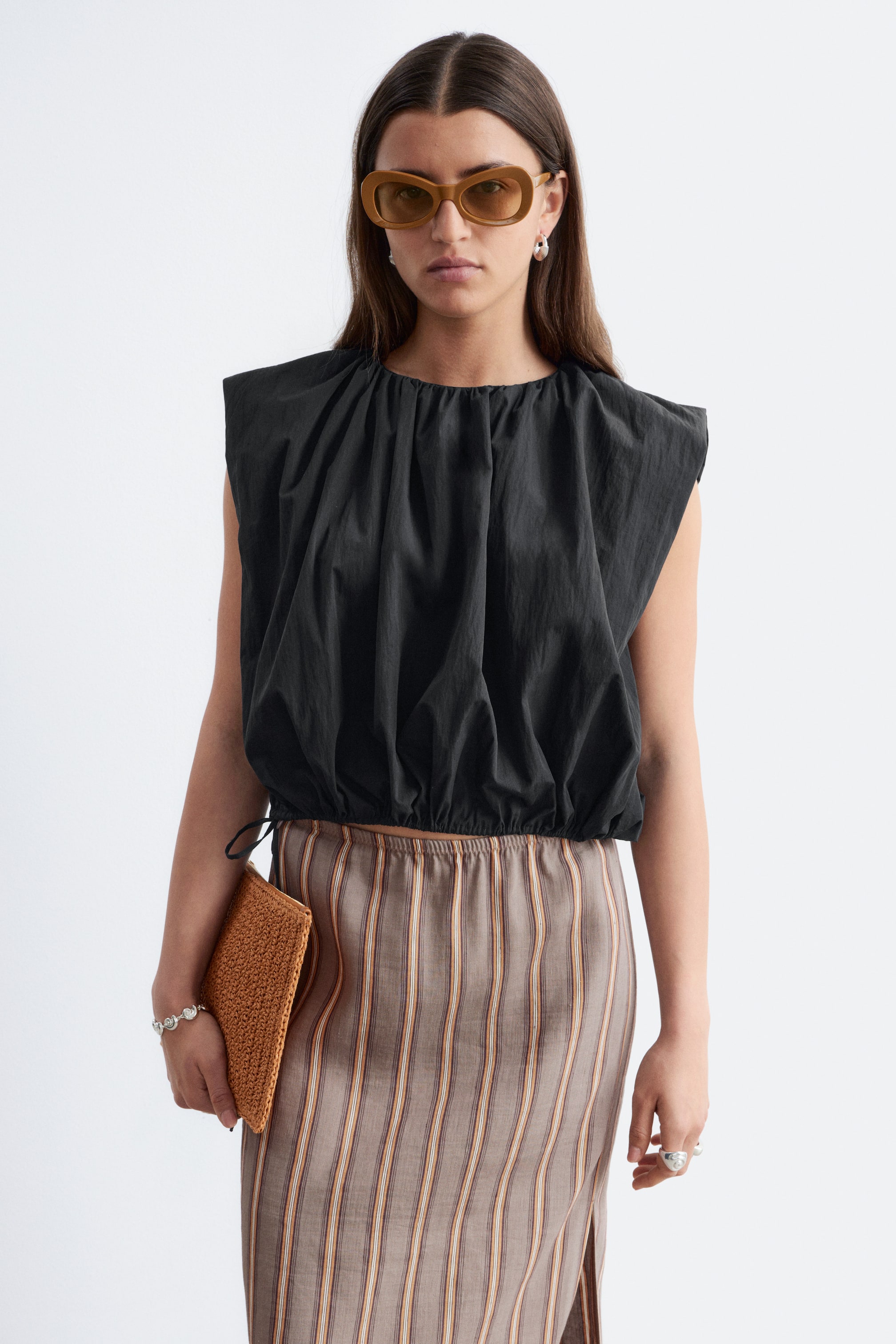 View larger image: The model is wearing a black gathered sleeveless top. It boasts a round neckline and an elasticated hem, and is styled with a striped midi skirt.