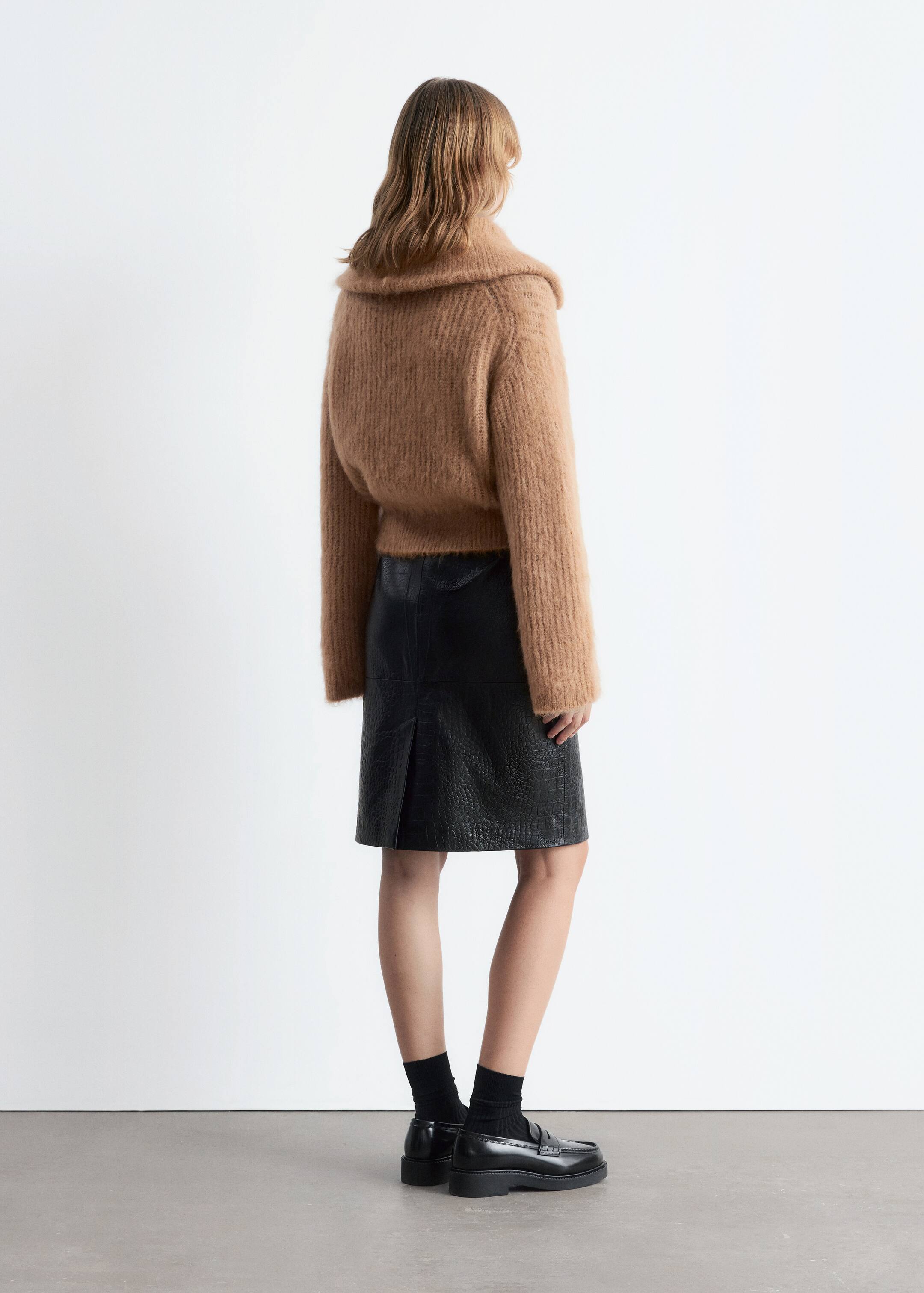 Grösseres Bild ansehen: Ein brauner Strickpullover mit flauschiger Textur und einem breiten, gerollten Kragen wird vom Model getragen. Der kurze Pullover wird über einem schwarzen, mit Krokoprägung versehenen Bleistiftrock mit Rückenschlitz gestylt, und schwarze Loafers werden getragen.