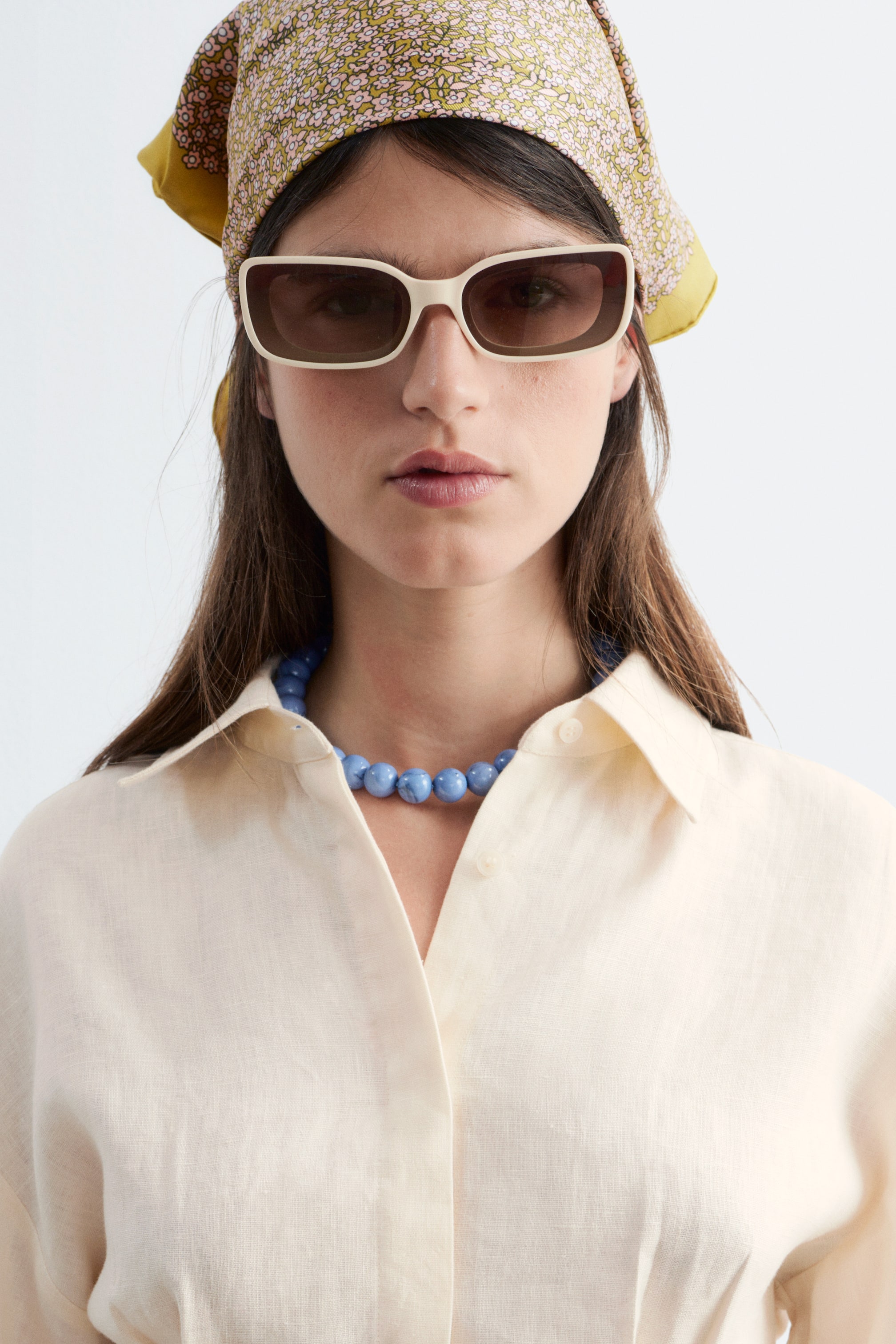 Agrandir l'image: Une chemise beige clair à manches longues, confectionnée en lin texturé, est dotée d'un col ouvert en pointe et d'une patte de boutonnage visible sur le devant, offrant une coupe décontractée. Un collier de perles bleues repose sur l'encolure de la chemise.