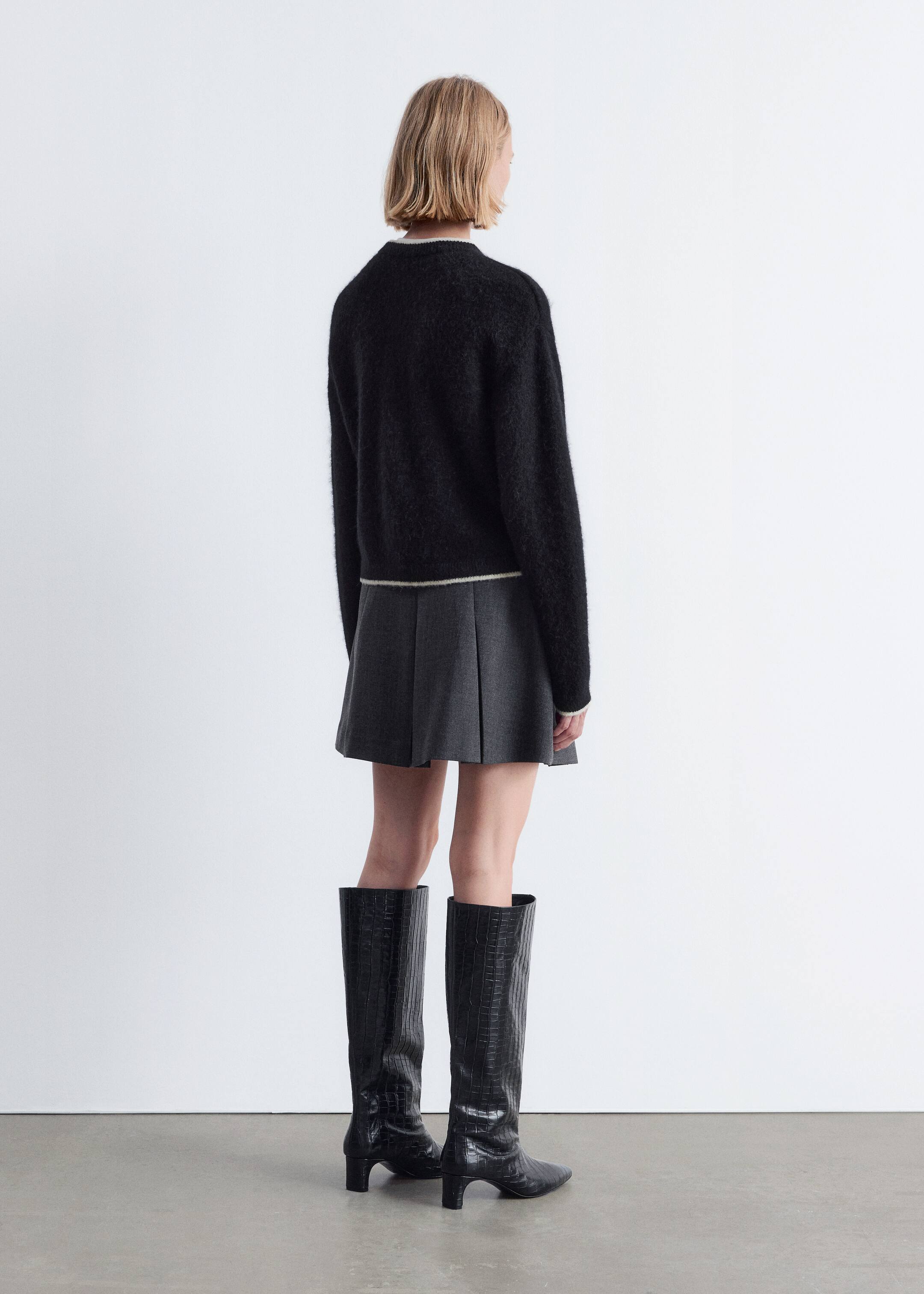 Agrandir l'image: Un pull noir en tricot duveteux avec une bande de couleur claire à l'ourlet est porté avec une mini-jupe plissée gris foncé et des bottes noires texturées jusqu'aux genoux, le tout vu de dos.