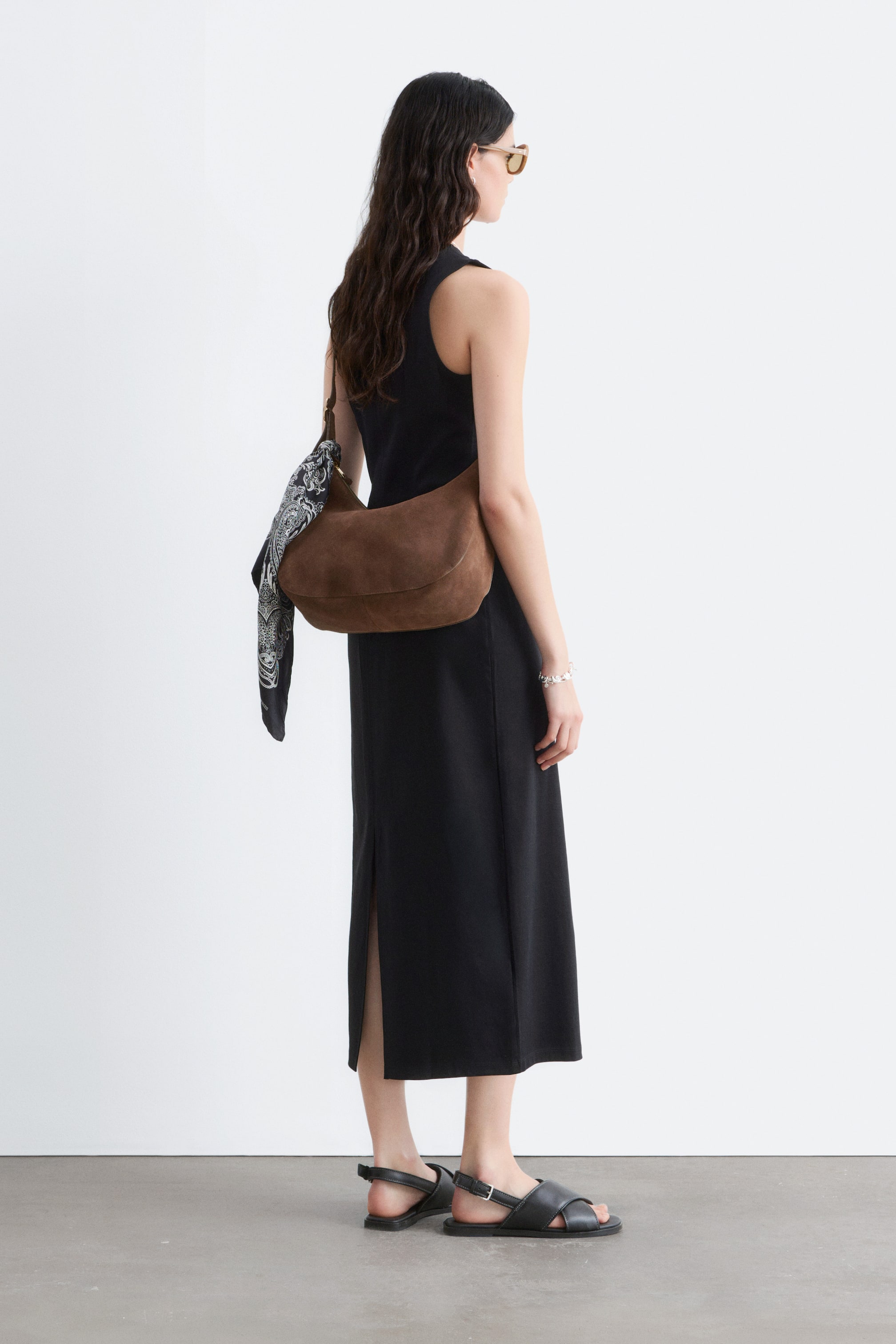 Agrandir l'image: La robe noire sans manches présente un col montant à l'arrière et une silhouette droite de longueur midi, avec une fente latérale visible sur le côté droit du porteur. Elle est montrée de dos et légèrement de biais.