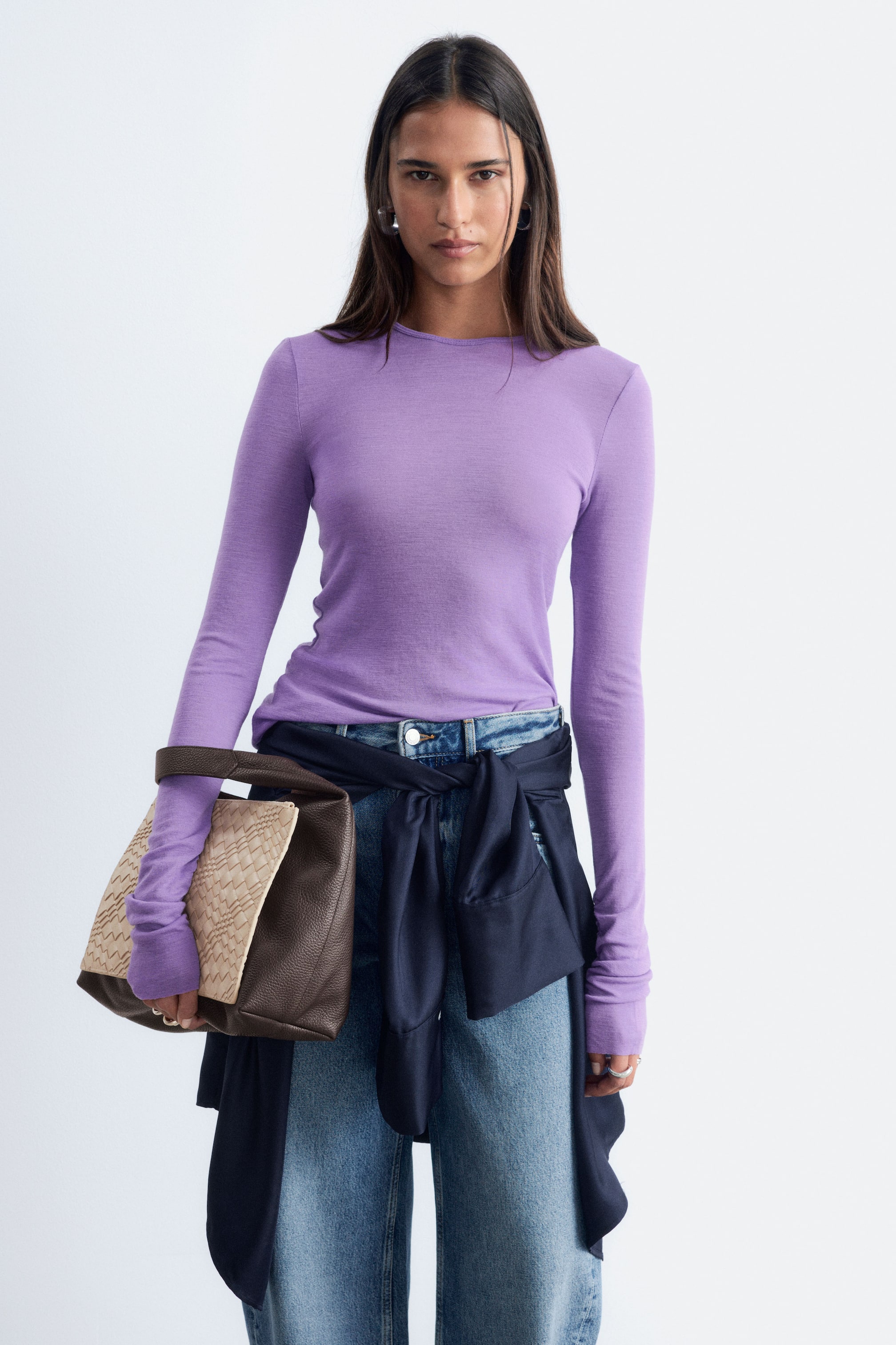 Ver imagem maior: A modelo usa uma camisola de lã roxa de malha lisa, com gola redonda e mangas compridas. A peça de corte justo está enfiada dentro de jeans de ganga azul, combinados com uma camisa escura atada à cintura.