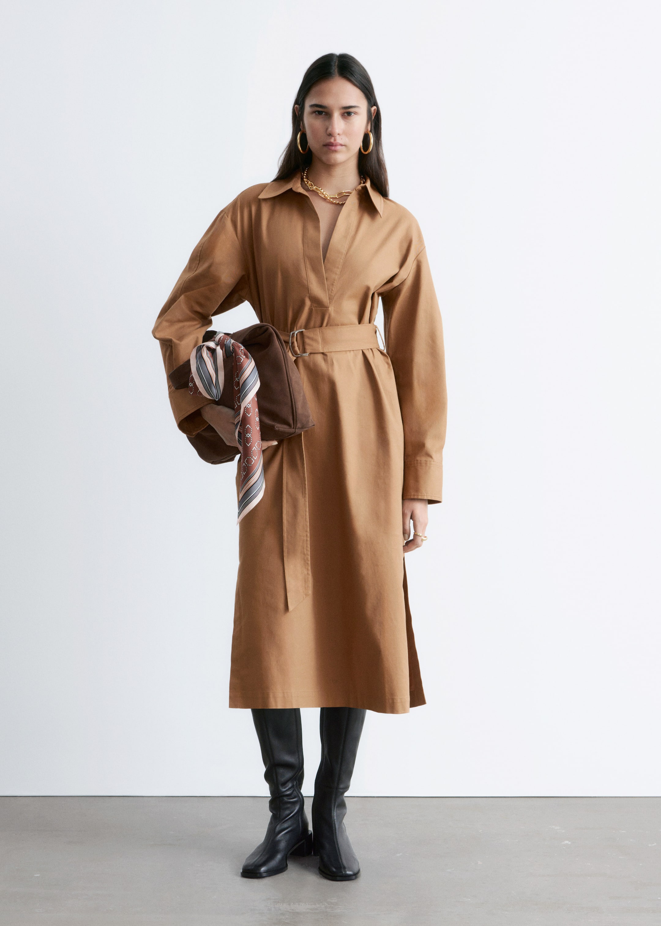 Größeres Bild anzeigen: Das sandfarbene Midikleid mit Kragen, V-förmigem Ausschnitt, langen Ärmeln und seitlichen Schlitzen am Saum wird vom Model getragen. Ein verstellbarer Taillengürtel betont die Taille des Kleides und schafft eine definierte Silhouette. Das Model trägt eine braune Tasche mit einem gemusterten Schal und kombiniert dazu schwarze Stiefel sowie goldenen Schmuck.