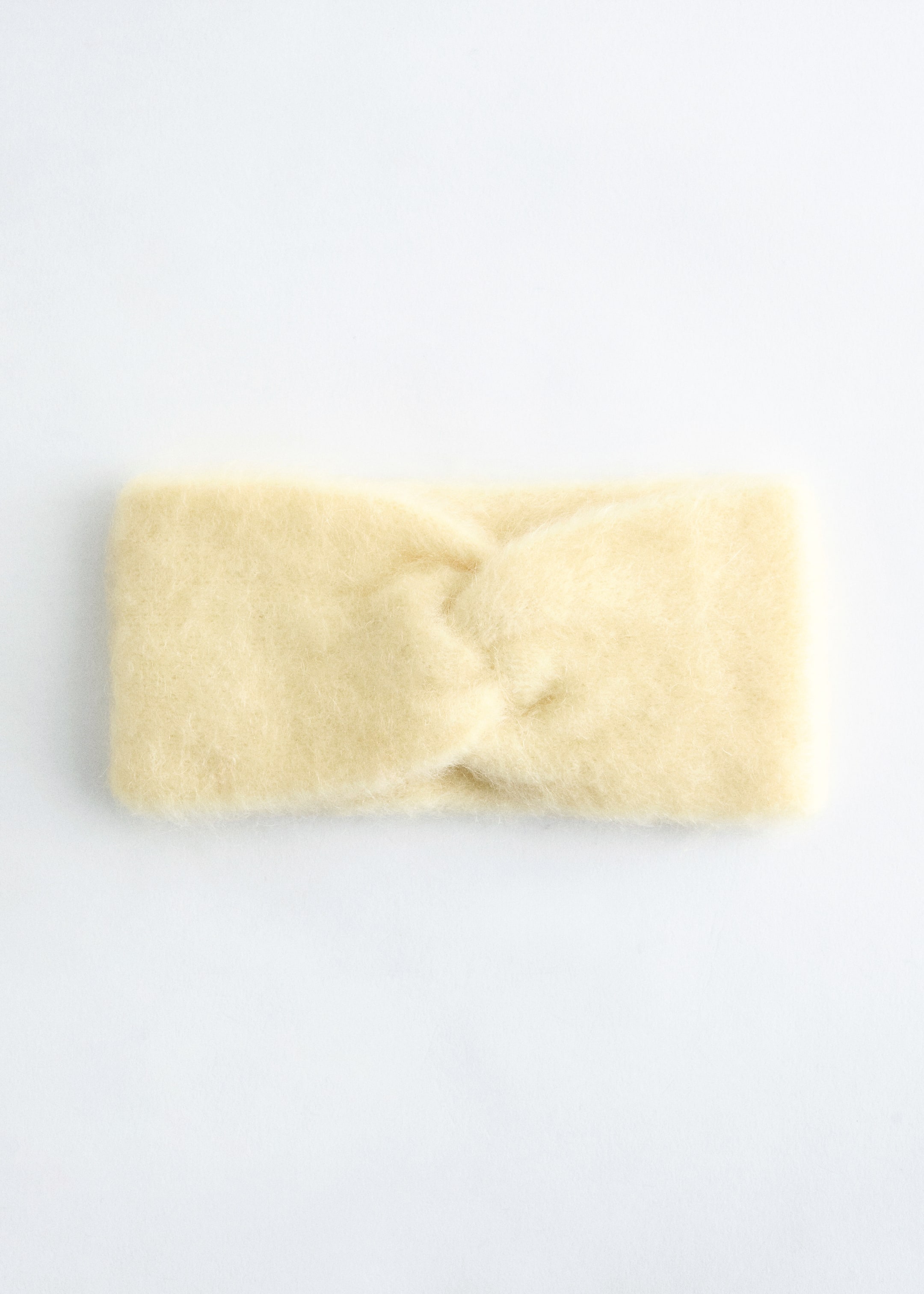 Grösseres Bild ansehen: Ein blassgelbes Stirnband aus Mohair-Mischung mit Twist-Front wird frontseitig präsentiert, mit einer sichtbar flauschigen Textur und einem markanten Twist-Detail in der Mitte.