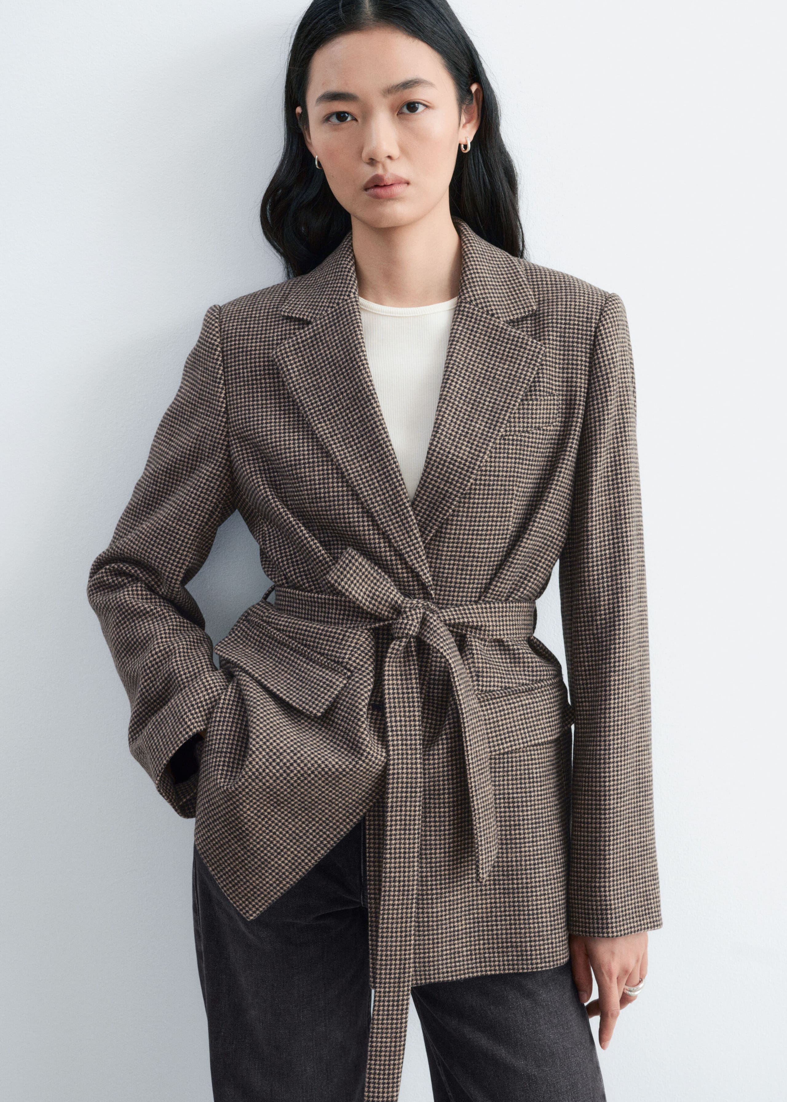 A brown houndstooth blazer with notched lapels is being worn by the model, featuring a matching fabric belt tied at the waist. The blazer is layered over a white crew-neck top and paired with dark grey trousers.