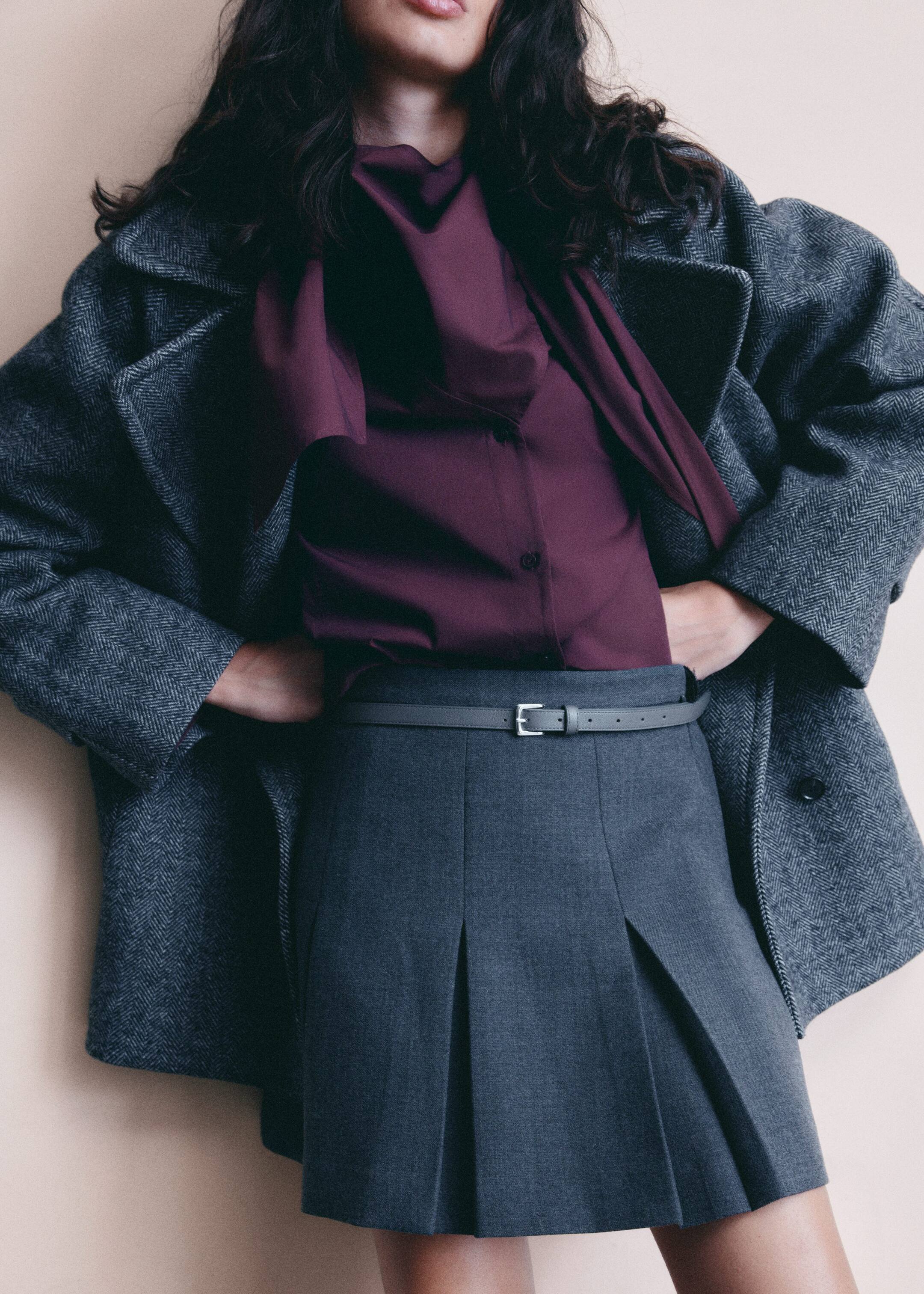 Agrandir l'image: Un manteau gris à chevrons est drapé sur un chemisier couleur prune avec un col drapé. Une mini-jupe plissée gris foncé est portée, serrée avec une fine ceinture grise ornée d'une boucle en argent.