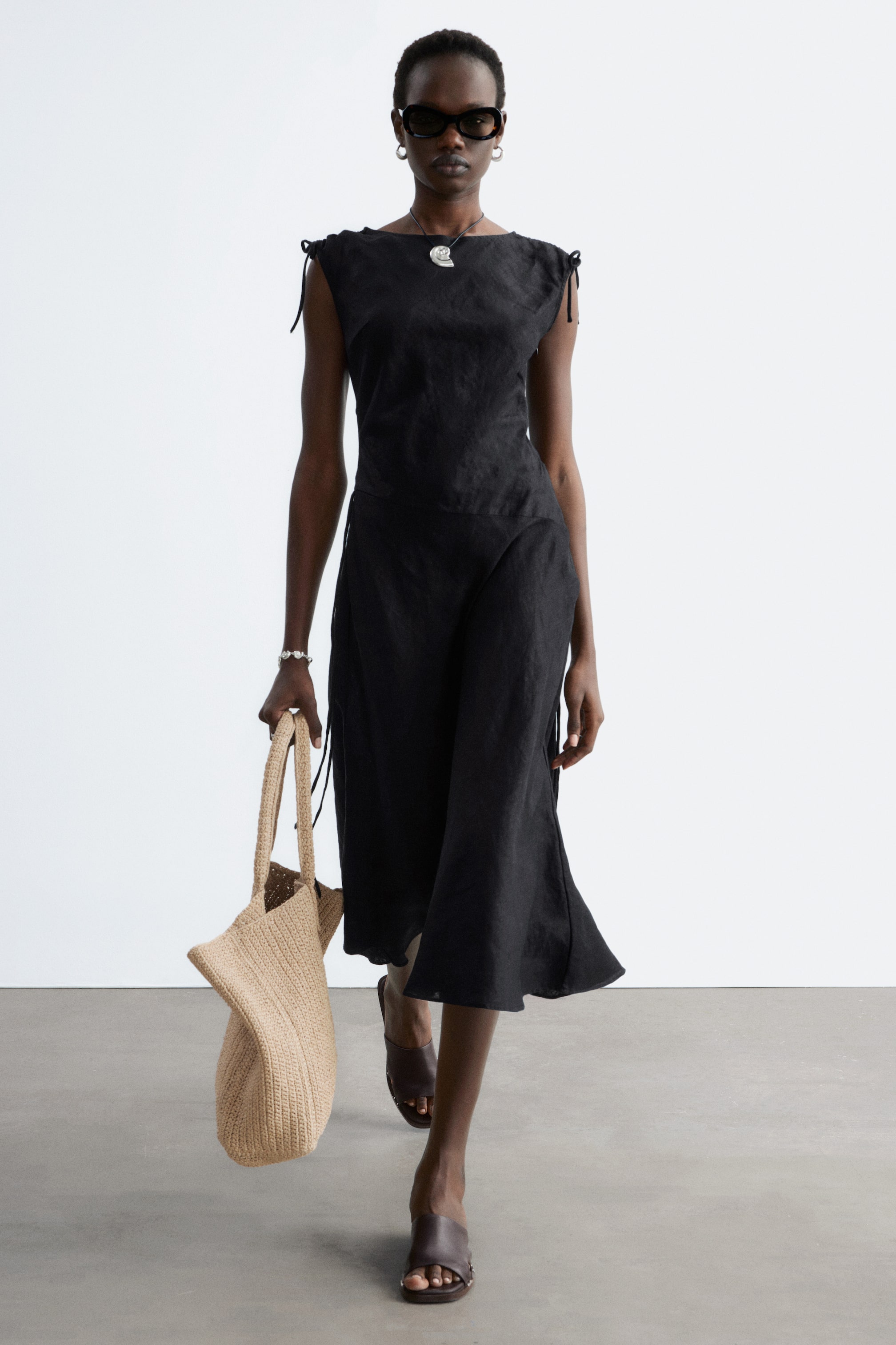 Ver imagem maior: Um vestido midi de linho preto apresenta um decote barco e laços nos ombros. O corpete ajustado flui para uma saia midi evasé com aberturas laterais.