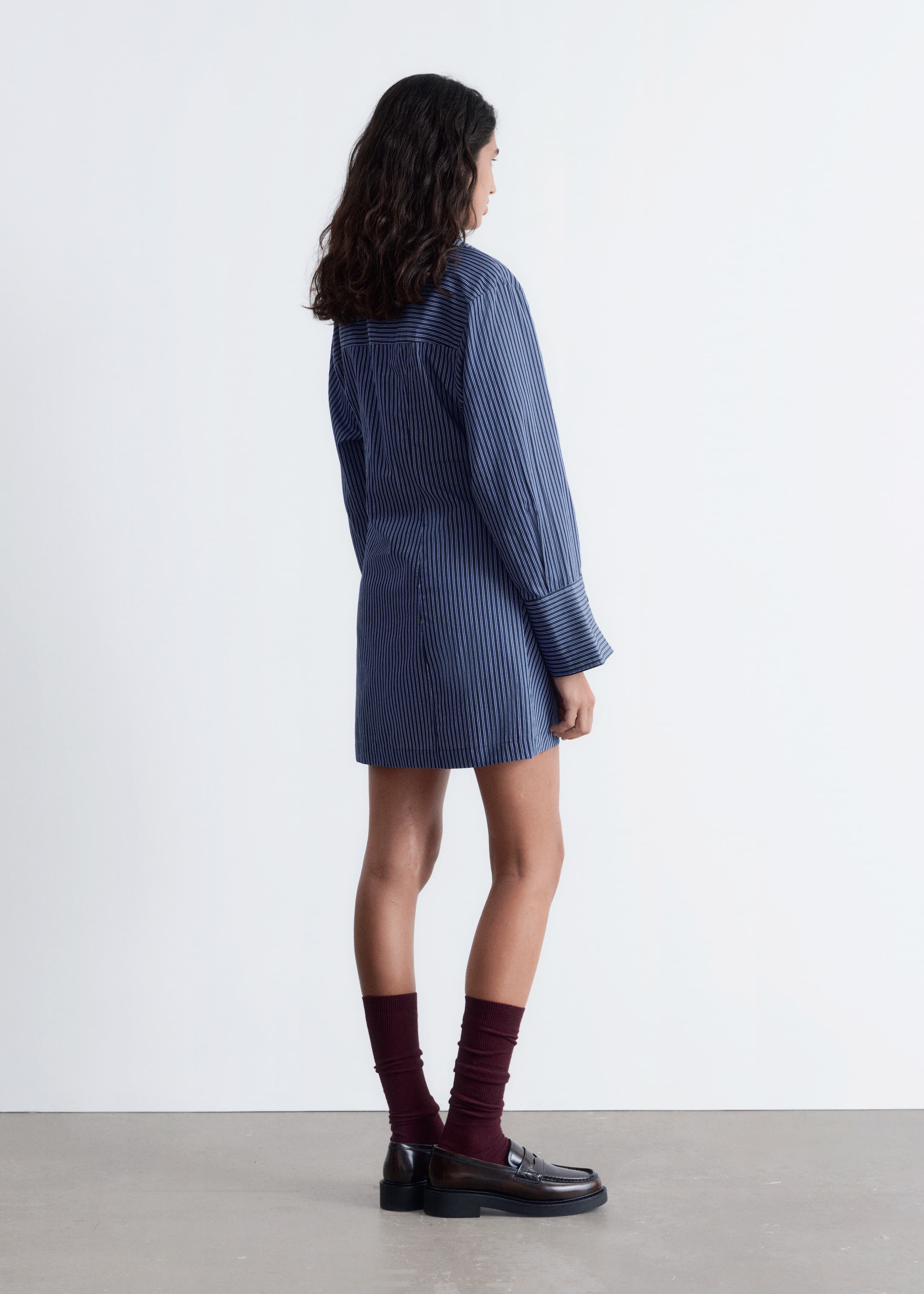 Visualizza immagine più grande: Un abito camicia a righe verticali blu è mostrato dal lato posteriore sinistro, caratterizzato da maniche lunghe con polsini larghi con bottoni e una vita stretta, che si estende fino a poco sopra il ginocchio.