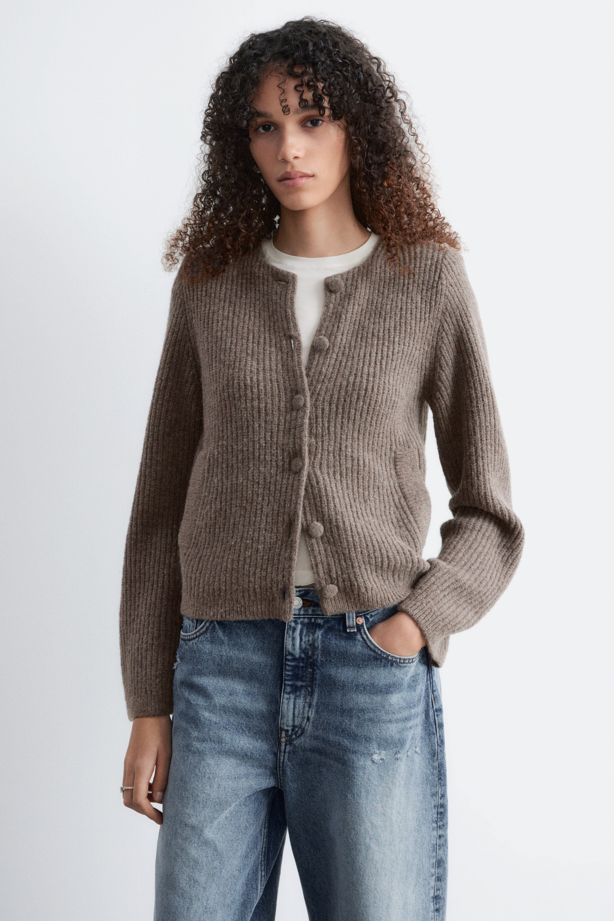 Se større billede: En brun ribstrikket cardigan bæres forfra, med en rund halsudskæring, en frontlukning med seks synlige runde knapper og lange ærmer, der afsluttes med ribbede manchetter, lagdelt over en hvid top.