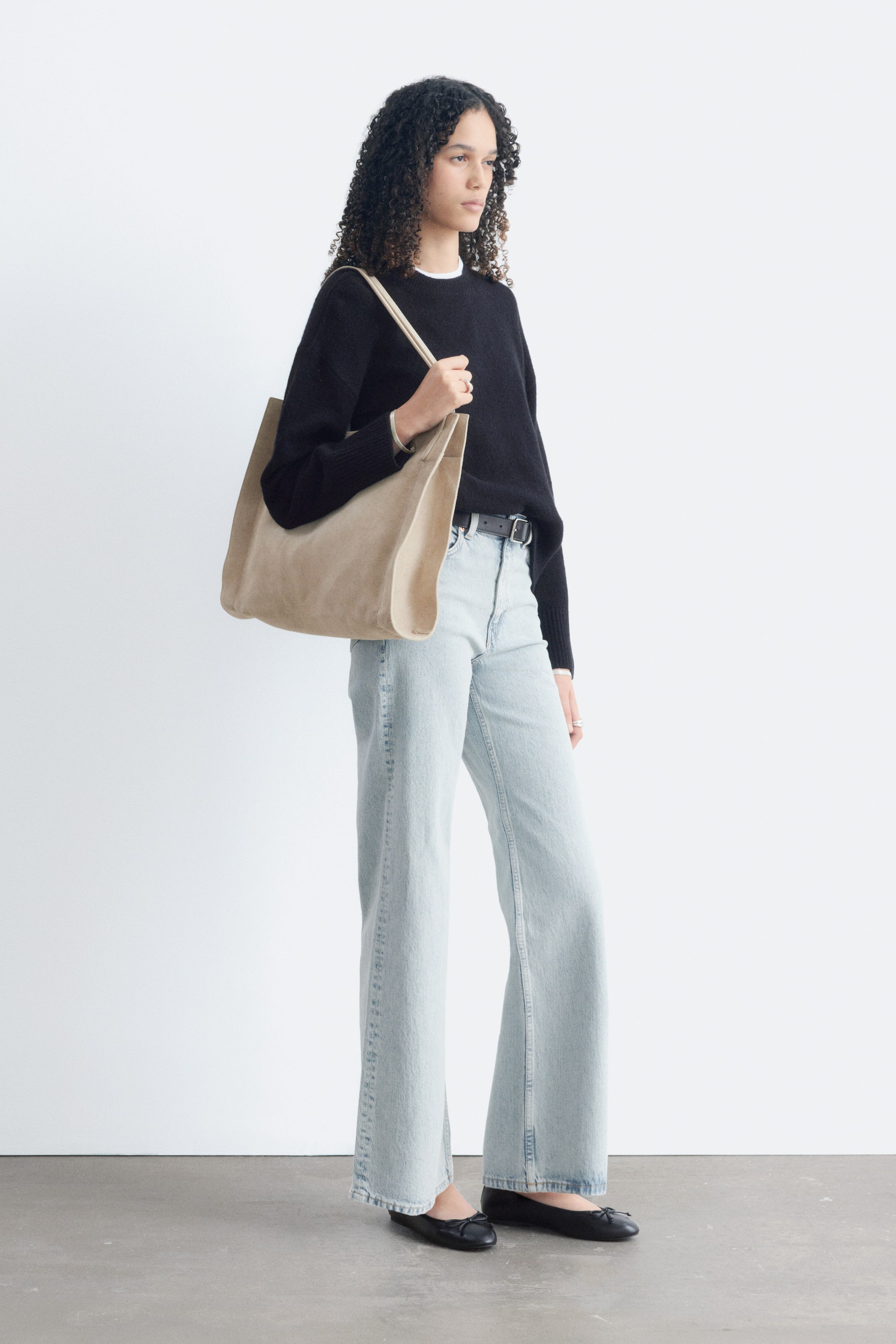 Visualizza immagine più grande: Una tote bag color tan chiaro è portata sulla spalla destra di una modella, rivolta verso l’osservatore. Presenta una forma rettangolare destrutturata e due manici superiori, ed è realizzata in un materiale morbido simile al suede.