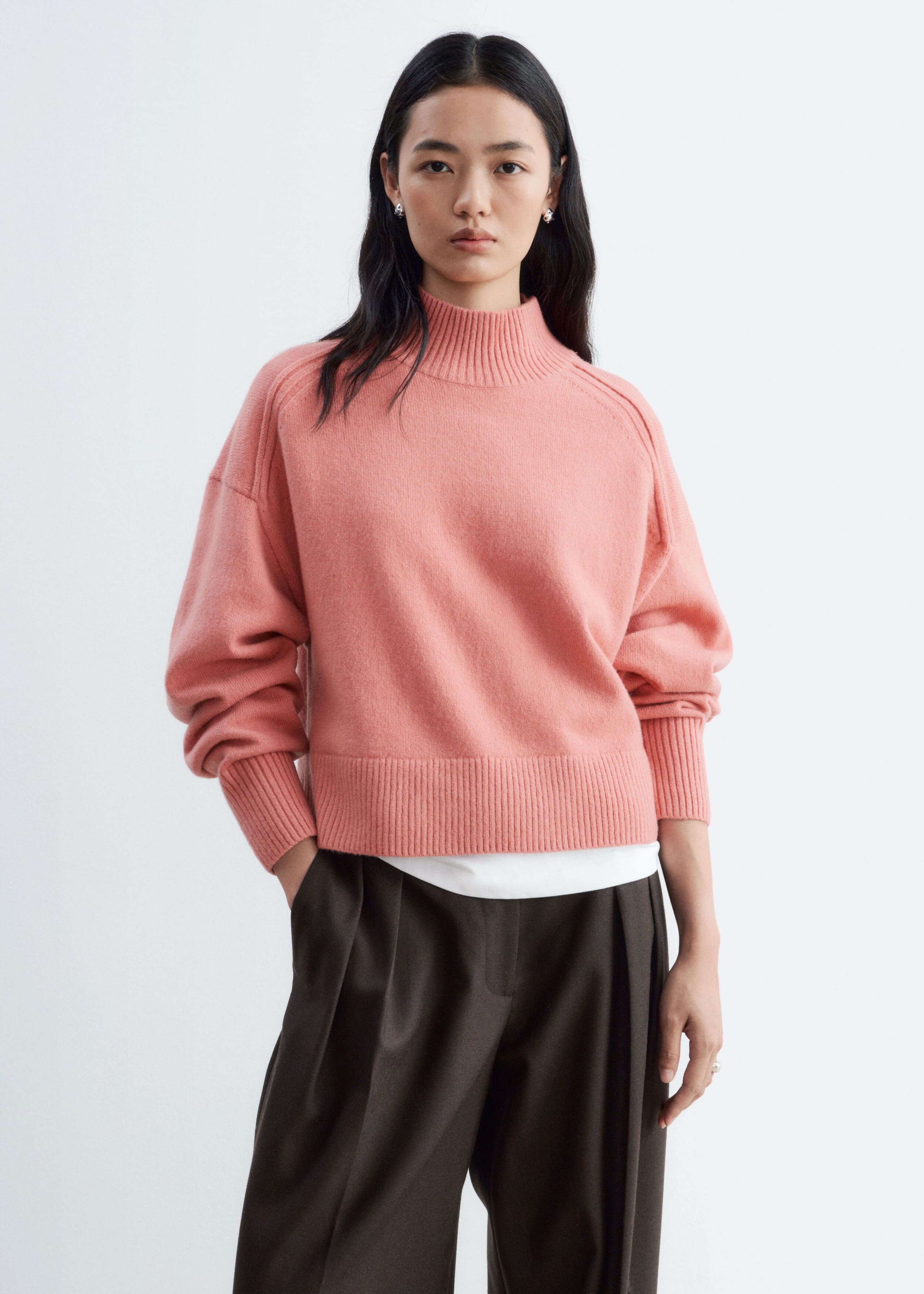 A pink mock neck sweater with forward-facing shoulder seams, ribbed cuffs, and a wide ribbed hem is being worn by the model. The knit sweater is layered over a white top and styled with dark brown pleated trousers.