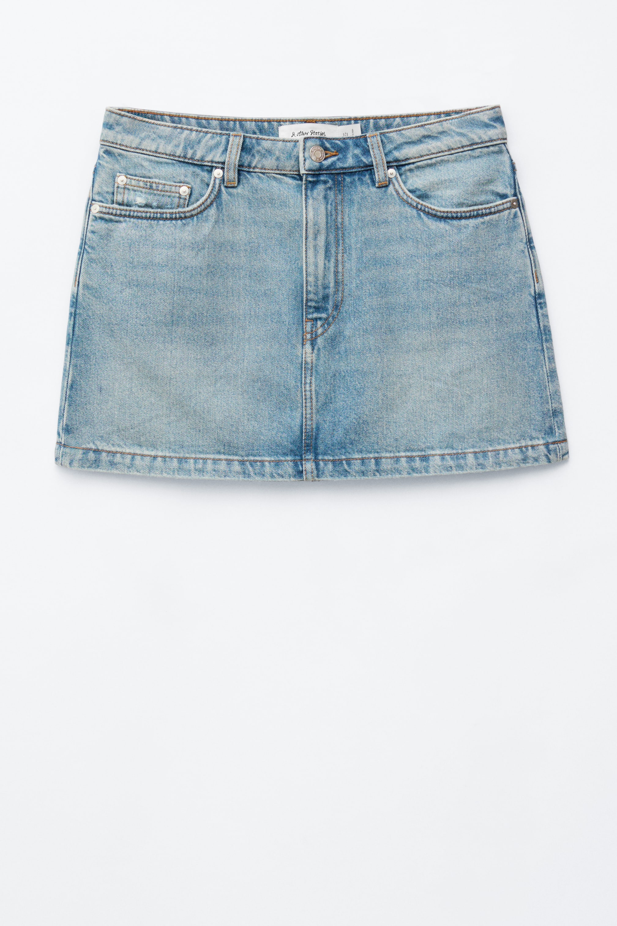 Ver imagem maior: Uma mini saia de ganga azul clara está a apresentar a sua parte frontal, com um clássico design de cinco bolsos com rebites de metal e fecho de botão. Apresenta um acabamento desbotado e usado.