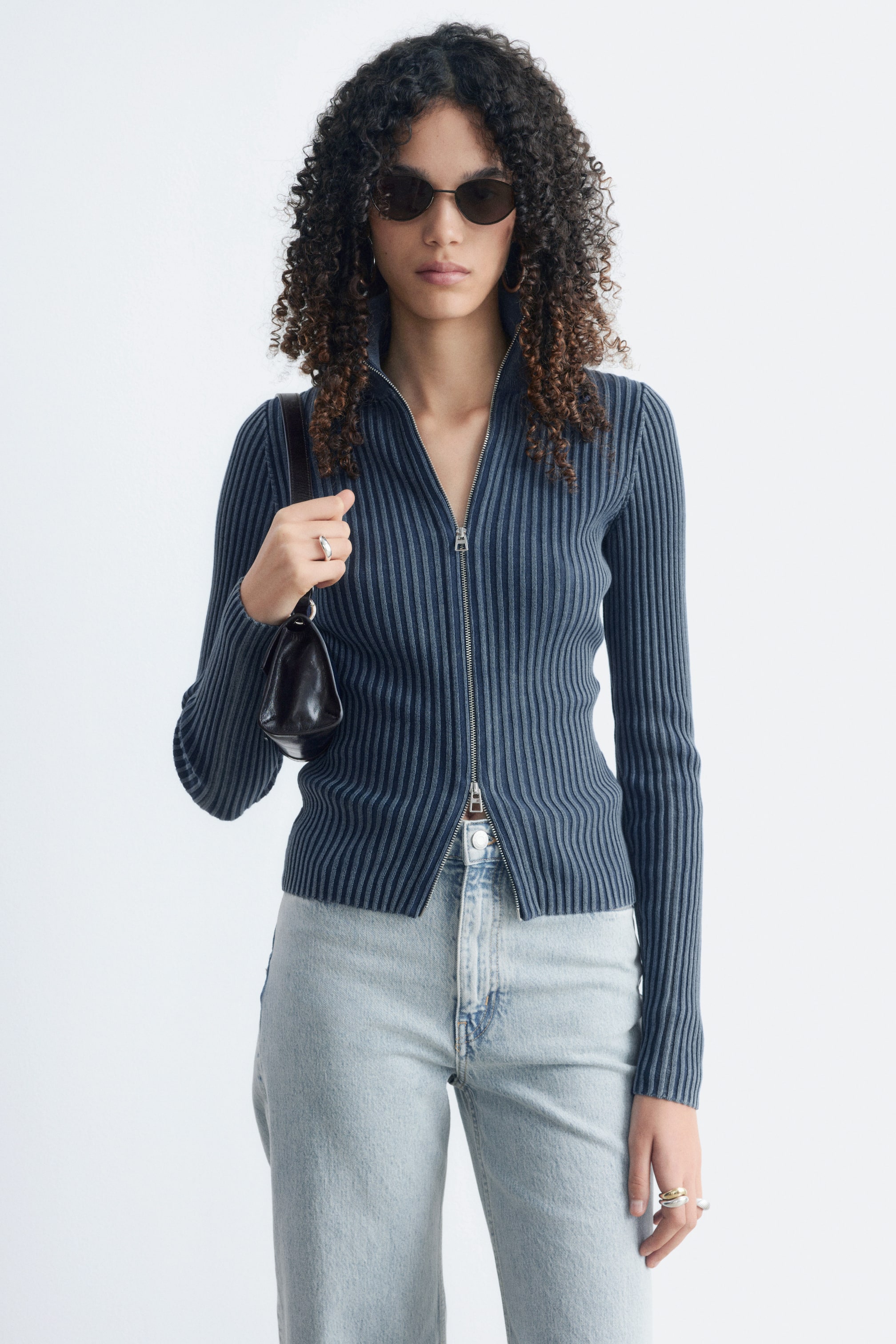 View larger image: The model is wearing a fitted dark blue long-sleeve cardigan with vertical ribbing, styled over light wash jeans. The cardigan features a stand collar with a two-way zip, partly open at the top and bottom.