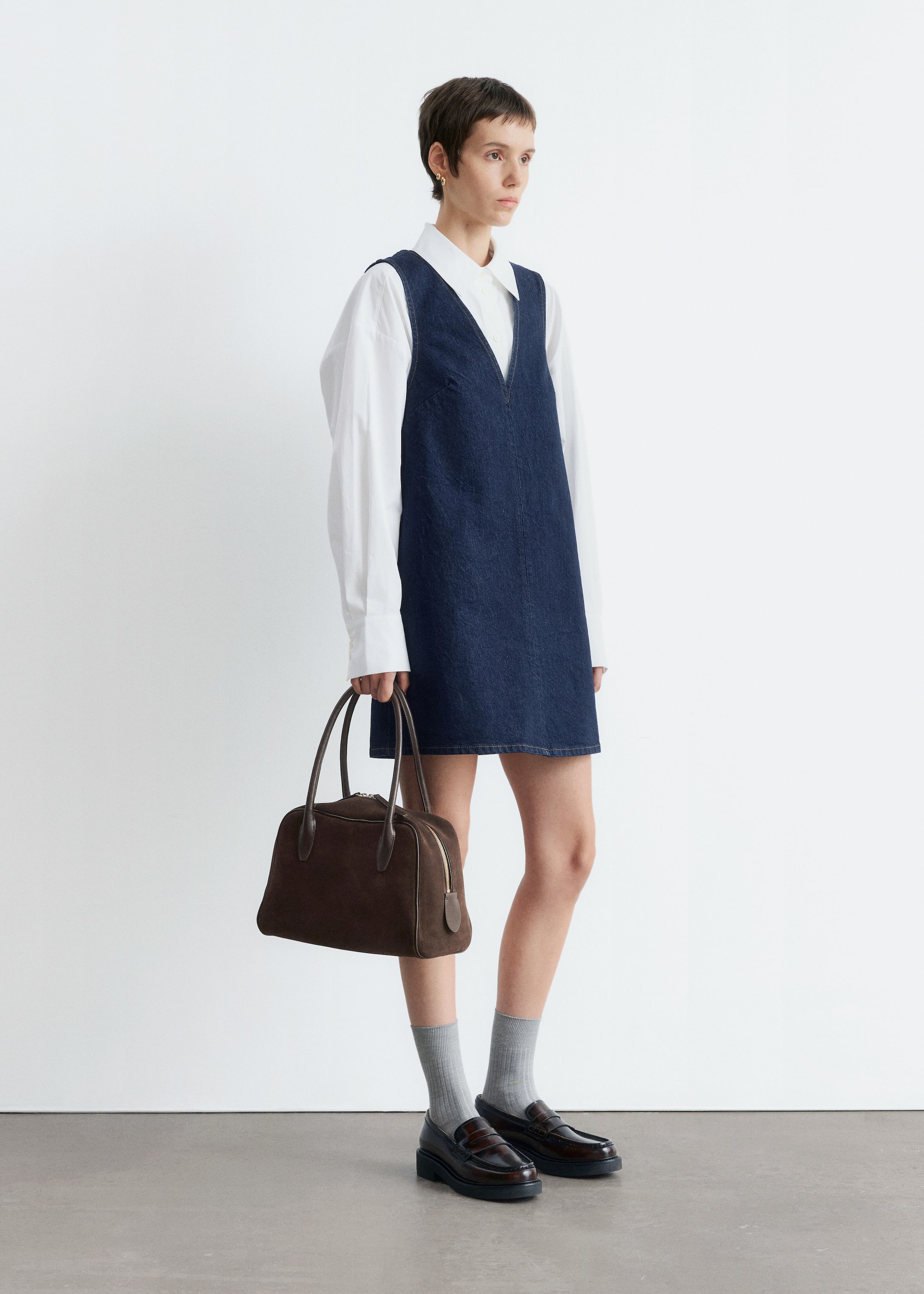 Ingrandisci l'immagine: Un mini abito in denim blu scuro, caratterizzato da una profonda scollatura a V e cuciture a contrasto visibili, è stratificato su una camicia bianca a maniche lunghe. Una borsa strutturata marrone scuro con una cerniera superiore è tenuta in mano, mentre mocassini marroni con suole spesse sono indossati con calzini grigi.