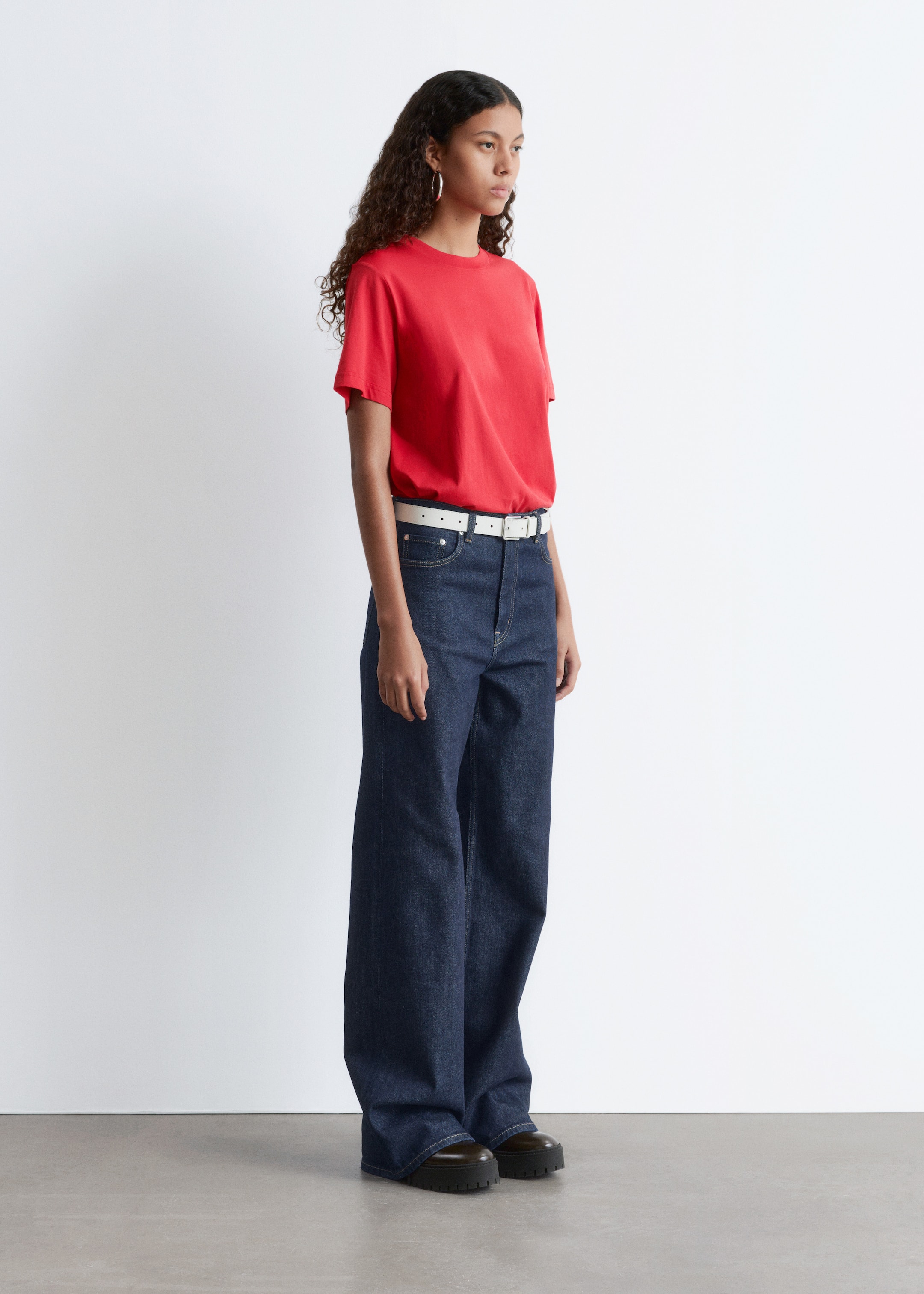 View larger image: A red short-sleeved crew neck t-shirt is being worn tucked into dark wash wide-leg jeans, which are being cinched by a white belt, with the full outfit being presented from a slight right-facing angle and completed by dark chunky-soled shoes.