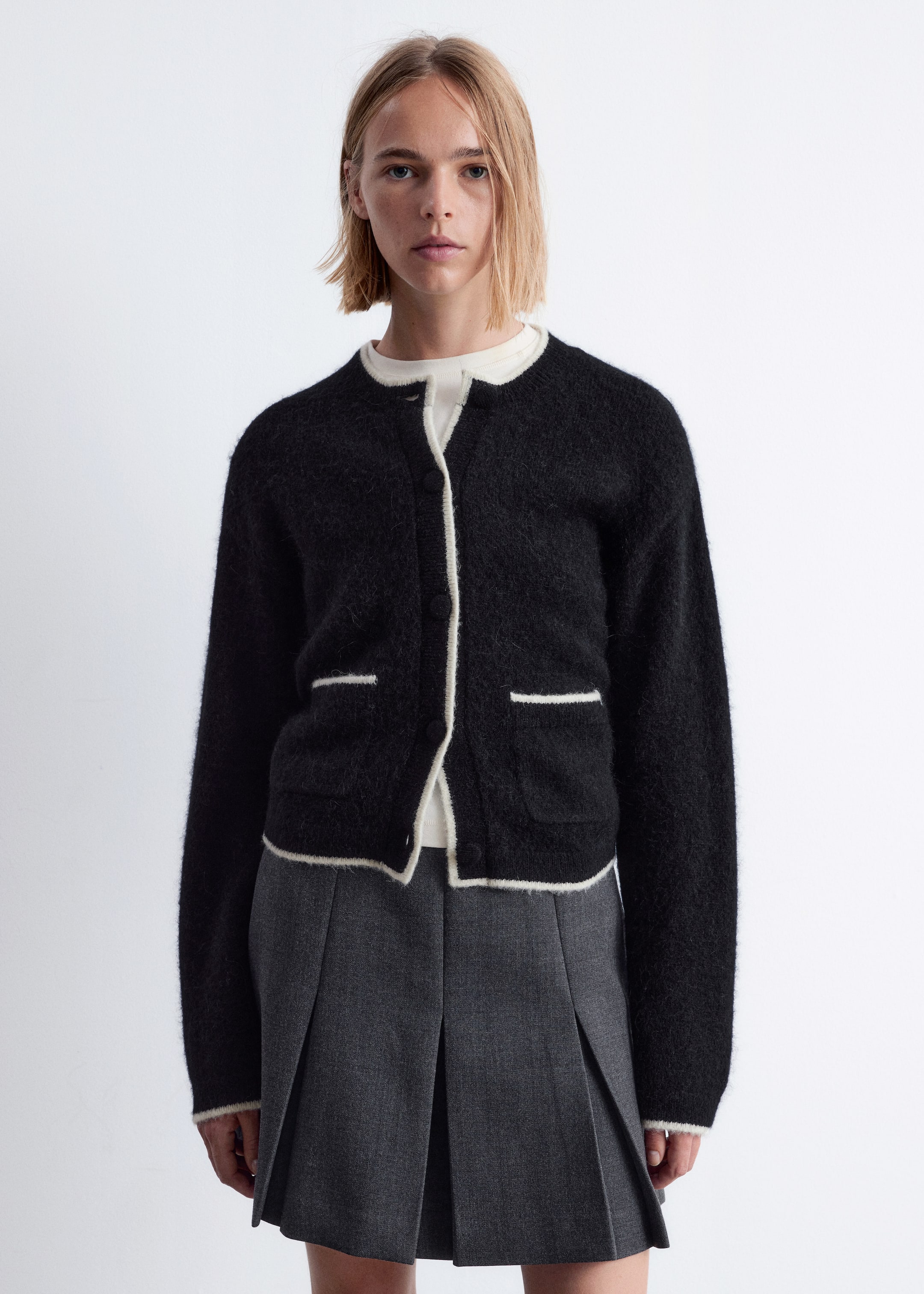Agrandir l'image: Un cardigan en tricot noir duveteux est présenté de face, avec un col rond, des manches longues et une bordure contrastante blanche le long du col, de l'ouverture avant, de l'ourlet et des poignets. Deux poches plaquées à l'avant, chacune avec une ligne de bordure blanche au-dessus de l'ouverture, sont visibles, et le cardigan est porté ouvert sur un haut de couleur claire.