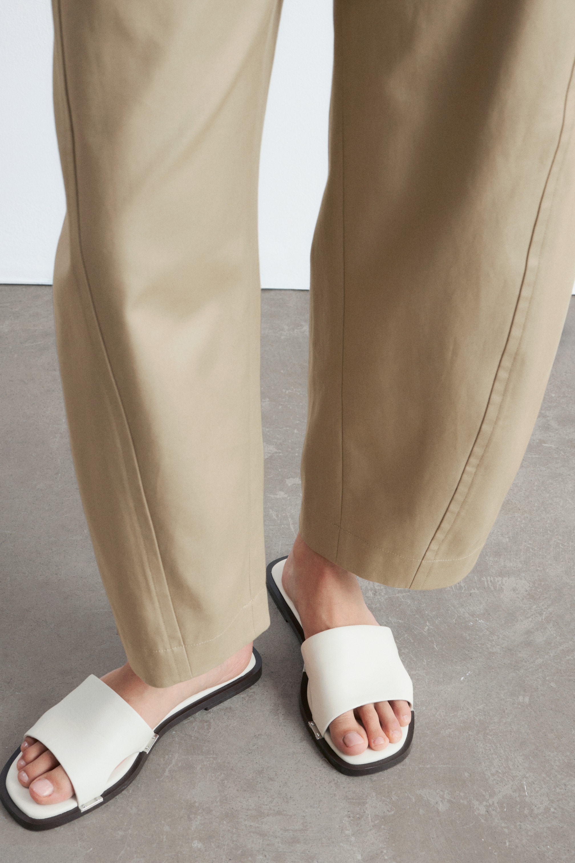 Se større billede: Beige ankellange bukser med snoede sidesømme, der danner en tøndeformet silhuet, bæres af modellen. Off-white læder slides med synlige metaldetaljer bæres også.