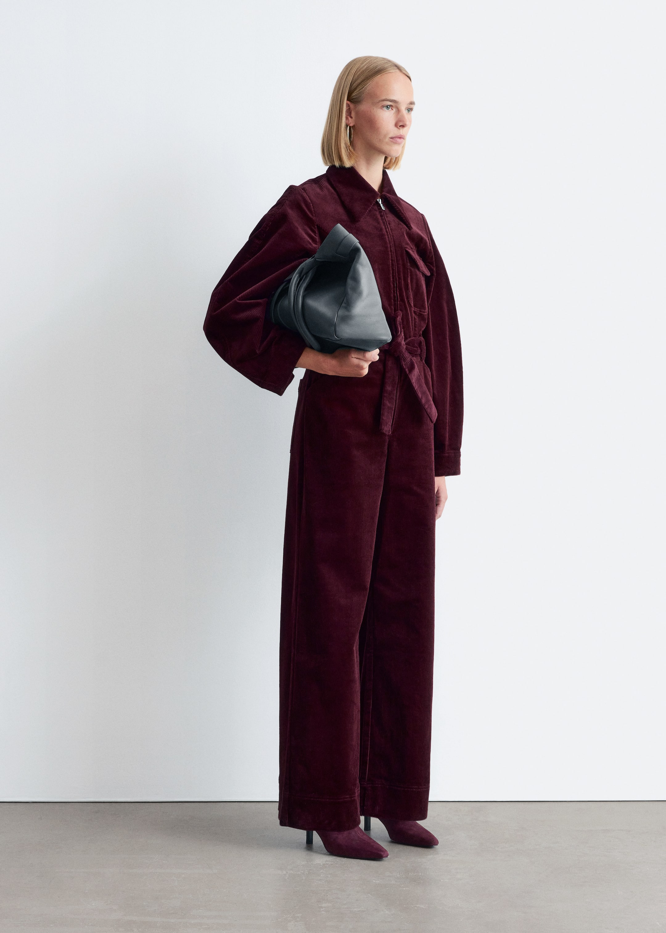 Größeres Bild anzeigen: Ein tief burgunderroter Cord-Jumpsuit wird getragen, aus einem leichten Winkel gesehen, mit einem Kragen, einem sichtbaren Reißverschluss vorne und langen voluminösen Ärmeln. Ein Selbstbindegürtel schnürt die Taille, und der Jumpsuit fließt in weite Hosenbeine. Eine dunkle, weich strukturierte Handtasche wird gehalten, und passende burgunderrote spitze Absätze werden getragen.