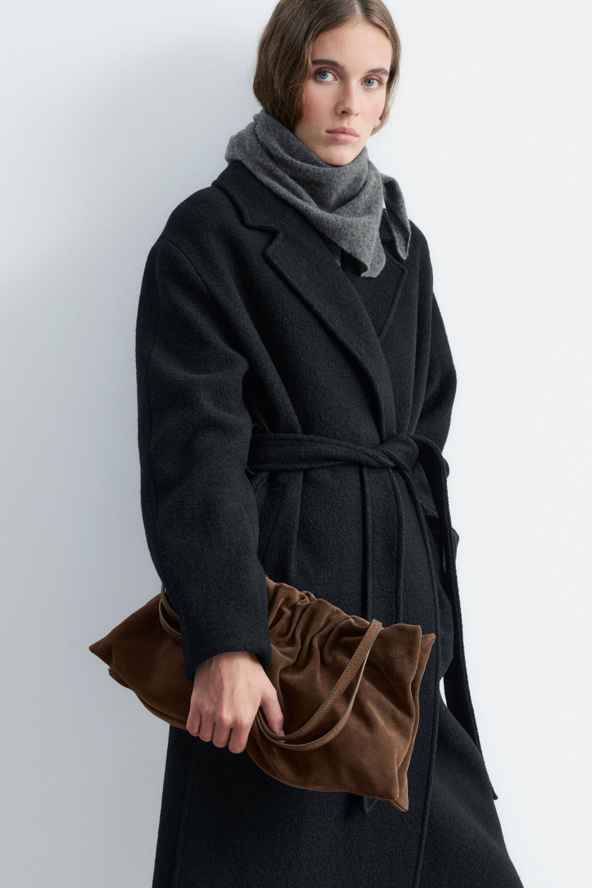 Visualizza immagine più grande: Una borsa a spalla marrone con effetto arricciato è portata dalla modella, che la tiene con una mano contro un cappotto nero con cintura. La borsa presenta una struttura morbida e raccolta, con sottili spallacci in pelle drappeggiati sulla mano.