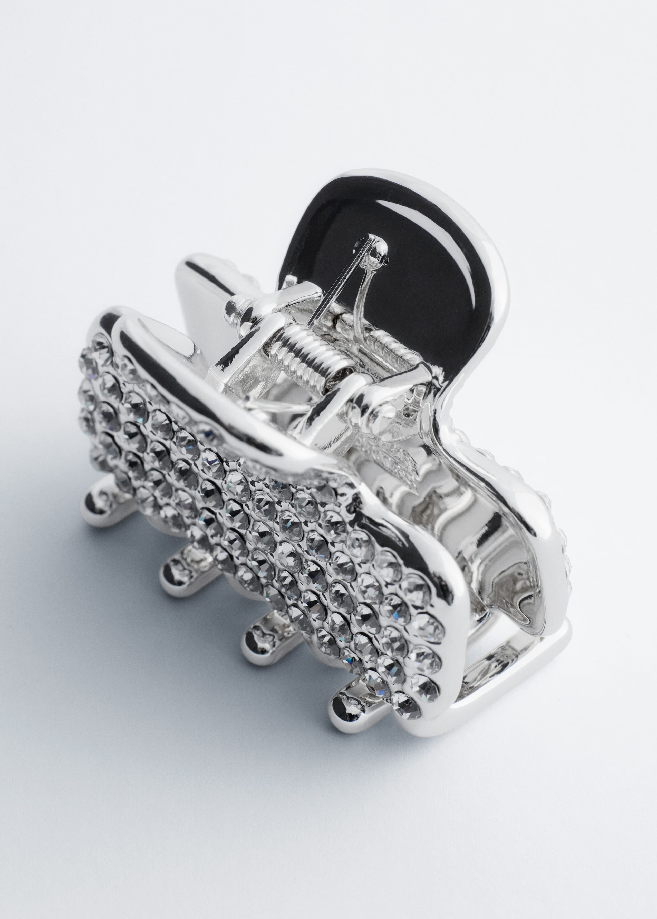 A silver-tone metal hair claw clip features an outer side densely encrusted with rows of clear, faceted rhinestones. The clip features a visible silver spring mechanism and a smooth, black-coated finger tab at the top.