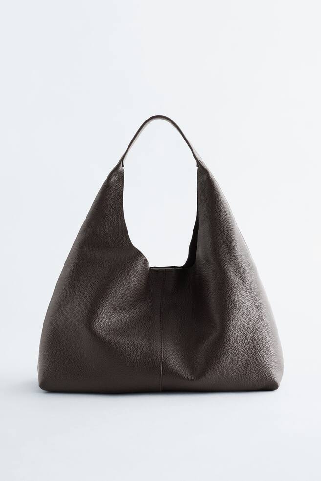 A dark brown, grained leather tote bag is shown from the front, featuring a soft, unstructured silhouette with a wide, integrated strap forming the top opening. - 1