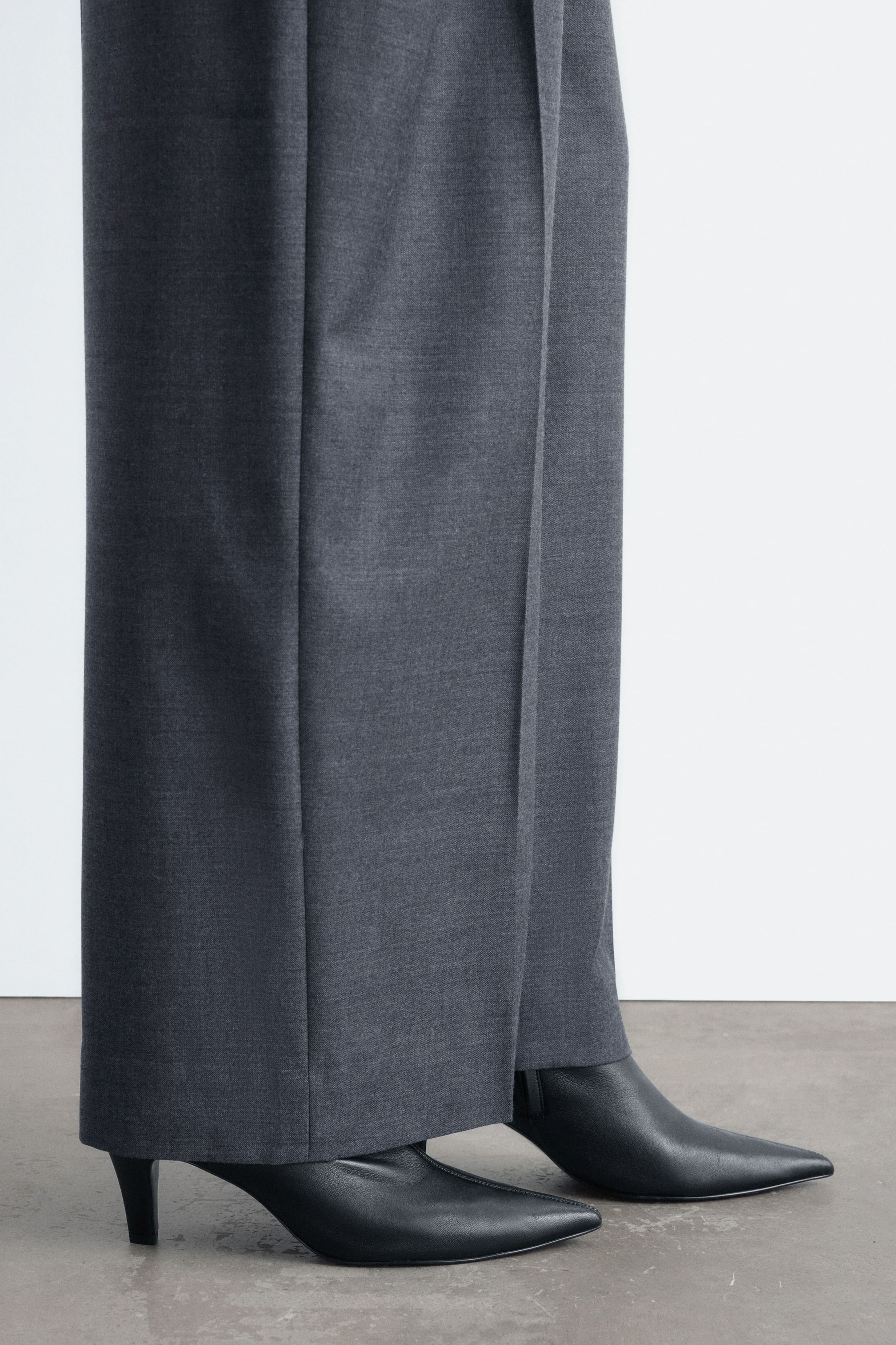 Grösseres Bild ansehen: Schwarze Sock-Boots aus Leder mit spitzer Zehenpartie und Stilettoabsatz werden vom Model unter weit geschnittenen grauen Hosen getragen.