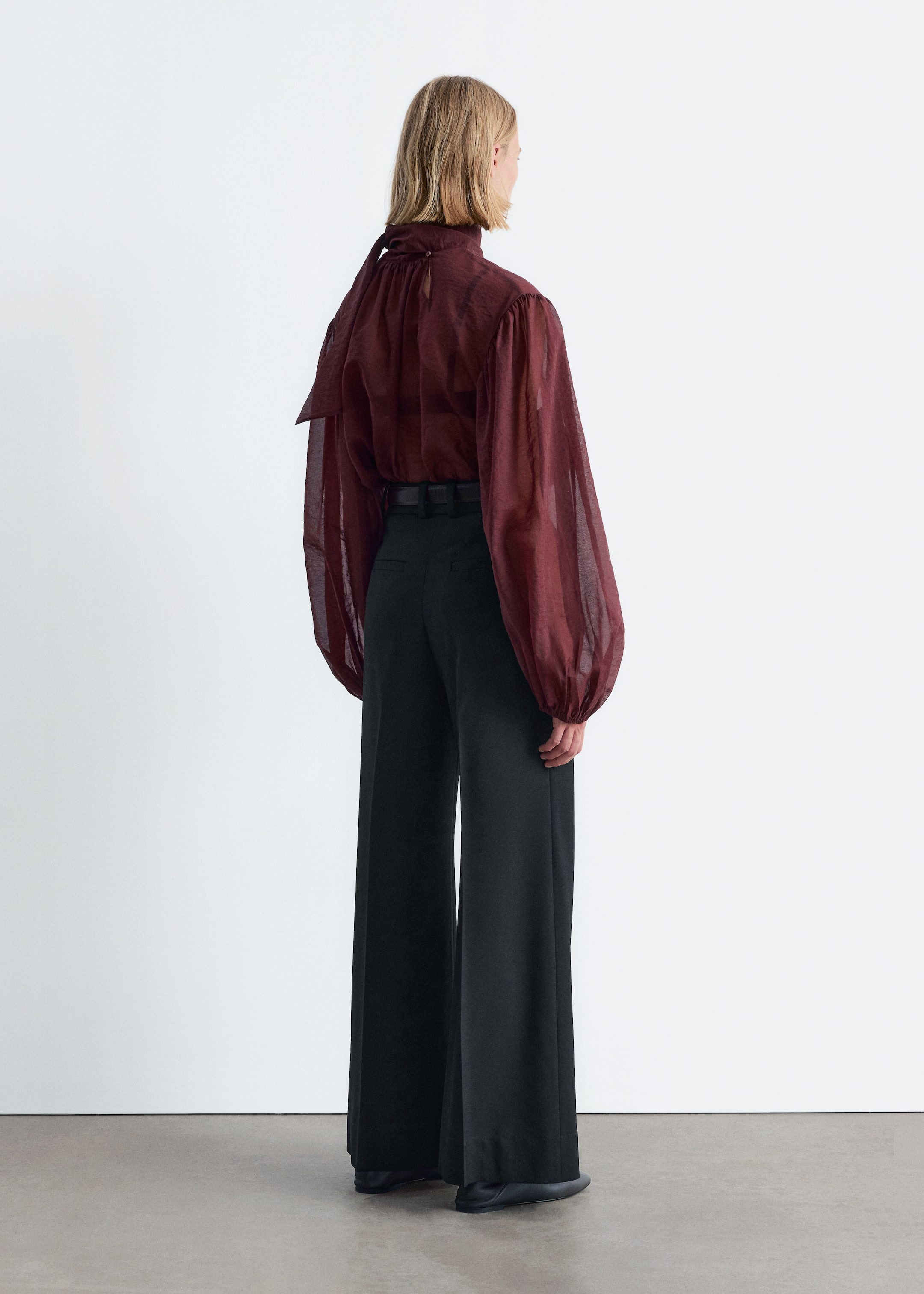 Visualizza immagine più grande: Una blusa trasparente di un rosso intenso viene presentata dal retro, caratterizzata da un collo alto con un dettaglio a fiocco e maniche lunghe voluminose raccolte ai polsini. La blusa è abbinata a pantaloni neri a gamba larga.