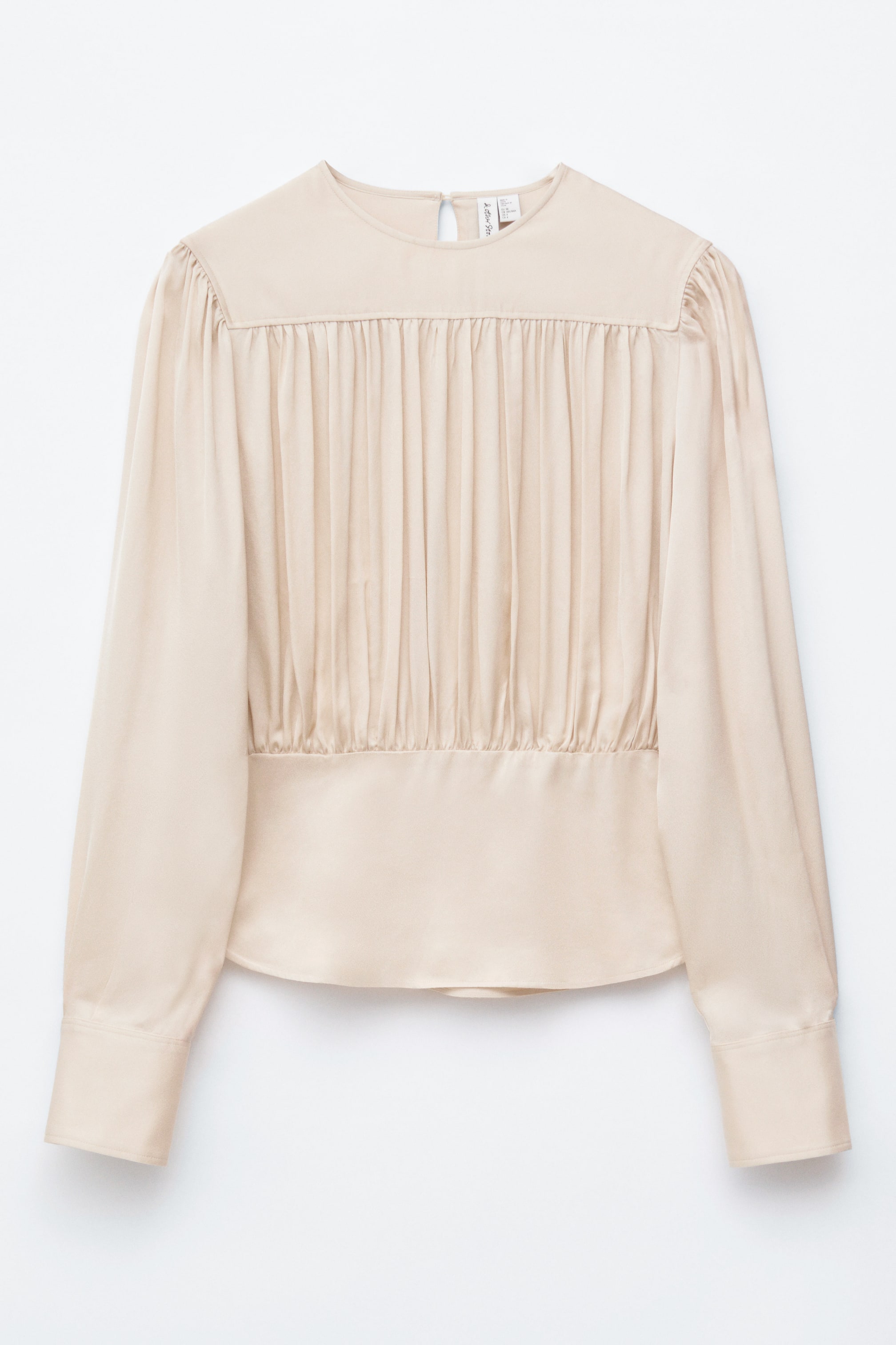 Zobacz większy obraz: Jasnobeżowa bluzka jest prezentowana z przodu, charakteryzująca się okrągłym dekoltem, długimi rękawami z mankietami zapinanymi na guziki oraz gładkim karczkiem na ramionach. Od karczku pionowe marszczenia tworzą efekt marszczenia na klatce piersiowej, zwężając się nad szeroką, gładką opaską dolną, wszystko to w gładkiej tkaninie z subtelnym połyskiem.