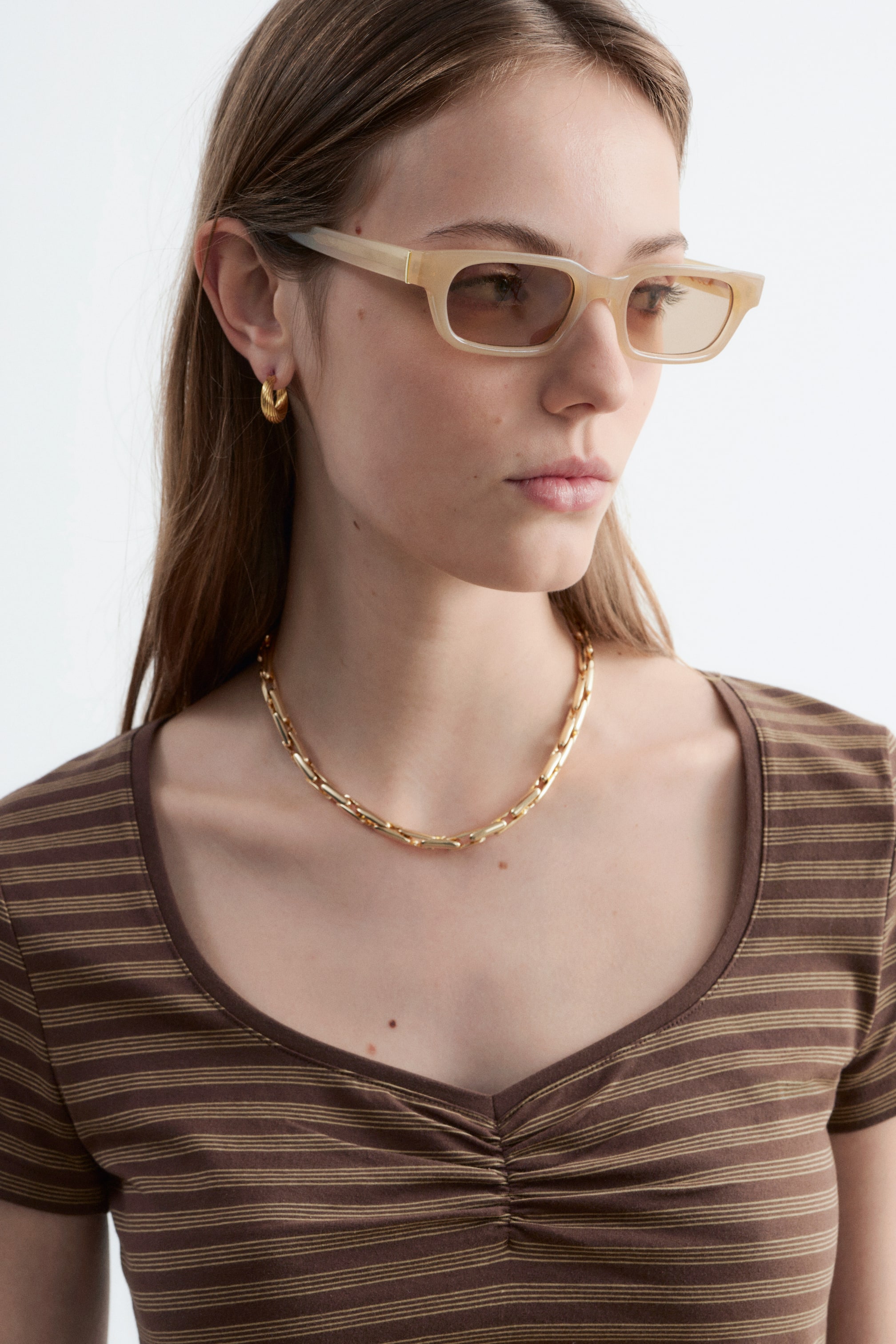 Zobacz większy obraz: Modelka ma na sobie brązowy, prążkowany, dopasowany top. Posiada dekolt w łódkę i marszczony biust, a na jego dekolcie spoczywa złoty naszyjnik.