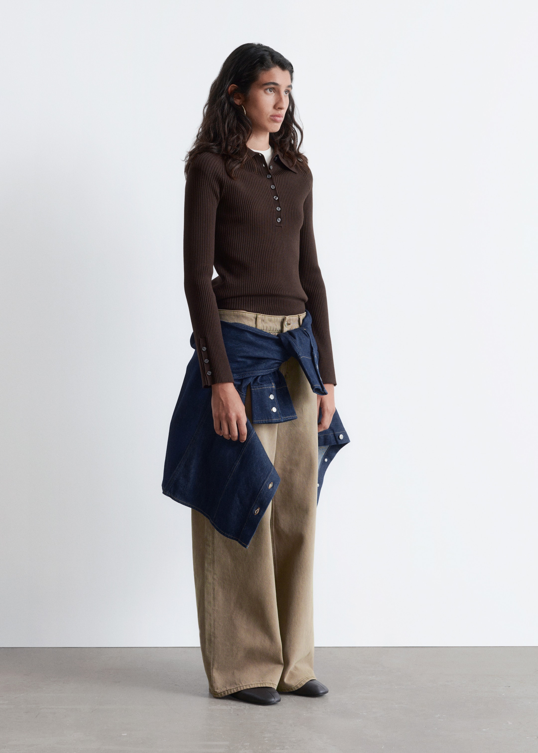 Grotere afbeelding bekijken: Een donkerbruine geribbelde gebreide poloshirt, met een klassieke kraag, een half knoopsluiting en mouwen met knoopmanchetten, wordt gedragen door het model. Het shirt wordt gelaagd over een wit ondershirt en gestyled met wijde beige broek en een spijkerjack dat om de taille is geknoopt.