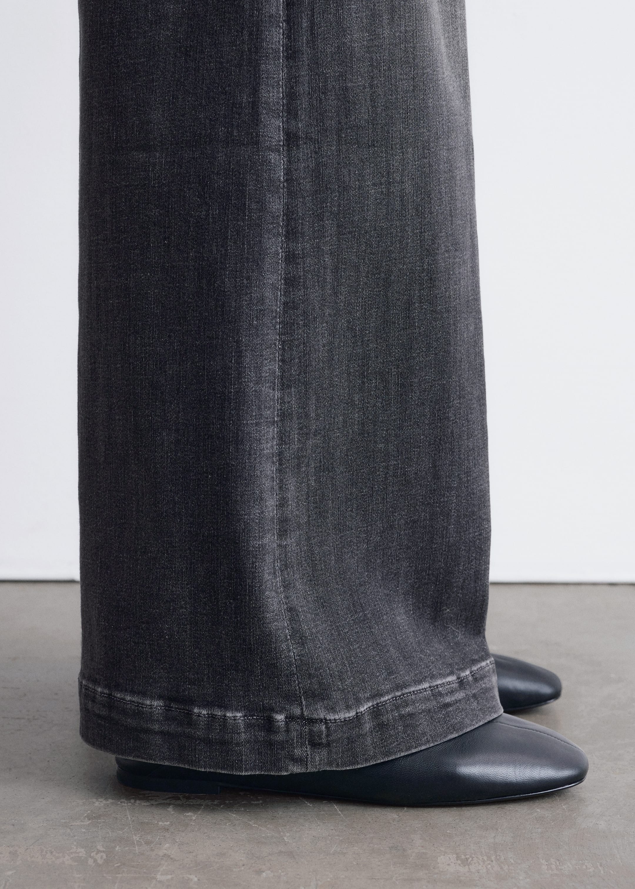 Visualizza immagine più grande: Pantaloni in denim grigio scuro a gamba larga sono mostrati da una vista laterale leggermente angolata. Una cucitura verticale prominente corre lungo la gamba, e il loro orlo ampio e leggermente scolorito poggia su scarpe nere piatte.