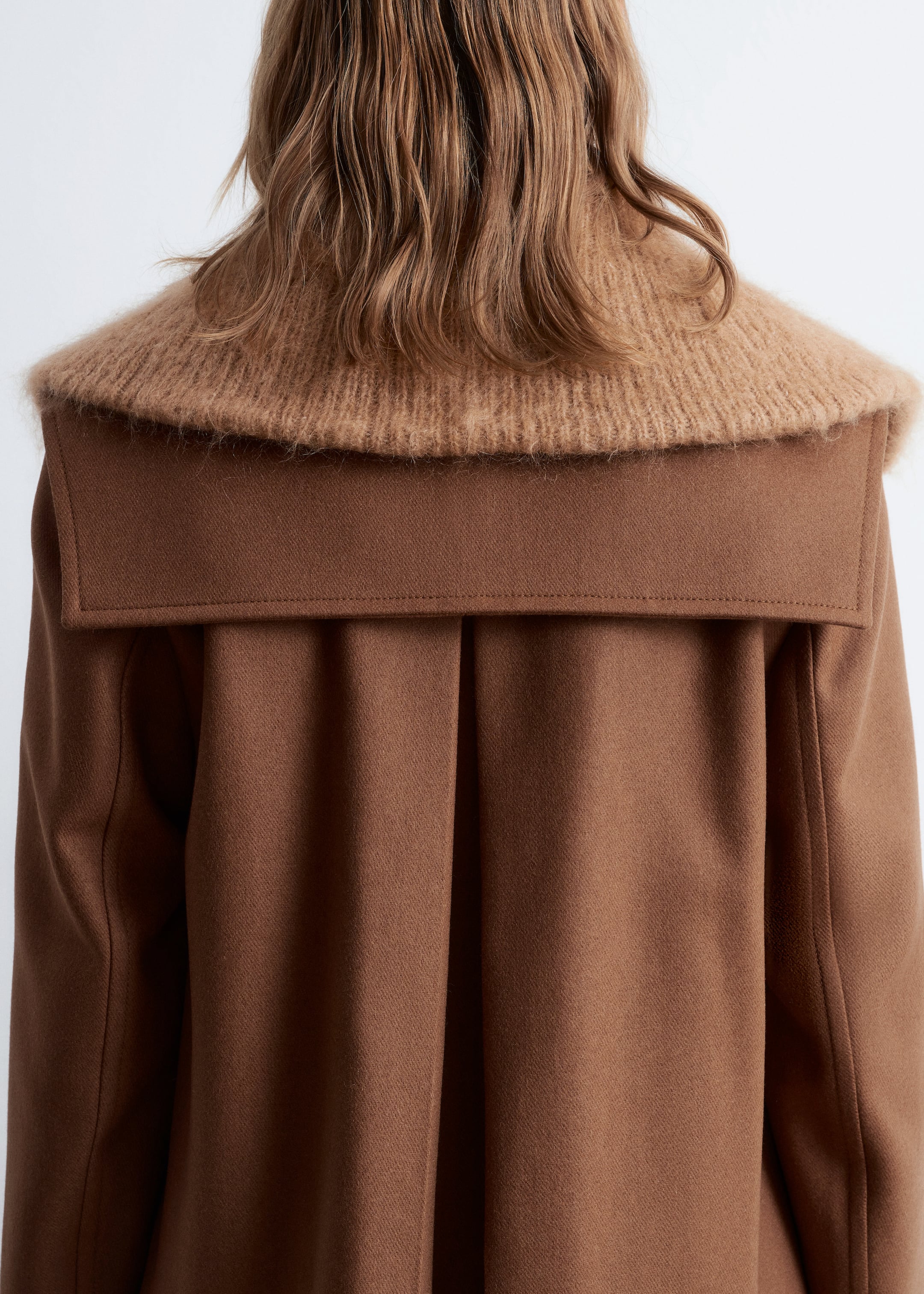 Grösseres Bild ansehen: Ein braune Jacke aus Wollmischung, die einen quadratischen Matrosenkragen am Rücken und eine Kellerfalte am hinteren Saum aufweist, wird vom Model getragen. Sie ist über einem hellbraunen, gerippten Strickkleidungsstück geschichtet, das über den Kragen der Jacke hinausreicht.