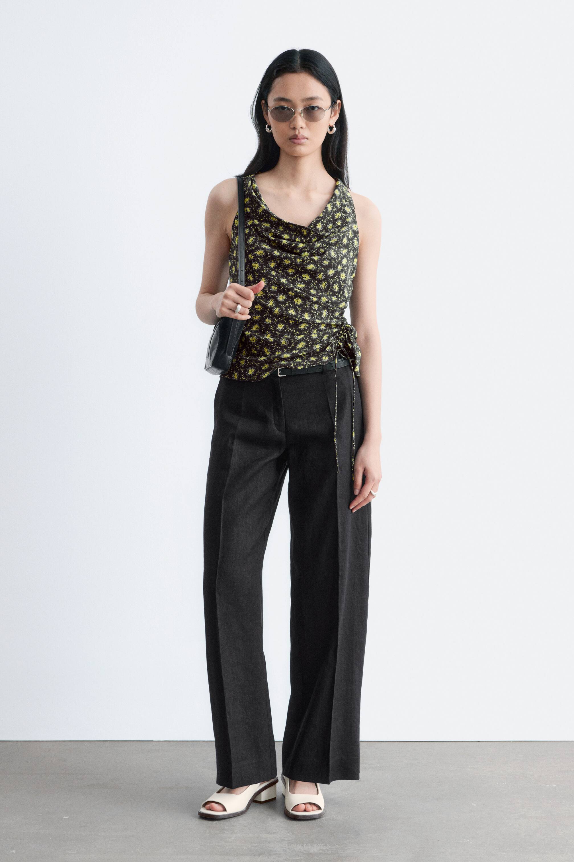 הצגת תמונה גדולה יותר: התלבושת כוללת חולצה ללא שרוולים עם צווארון קפלים ובסיס שחור עם דוגמת פרחים צהובים, המציגה קשירה עם שרוך שיוצרת כיווצים בצד השמאלי. מכנסיים כהים רחבים מציגים קפל קדמי חד, מאובטחים על ידי חגורה שחורה דקה, בעוד שסנדלים בצבע קרם עם עקבים בלוק פתוחים משלימים את המראה.