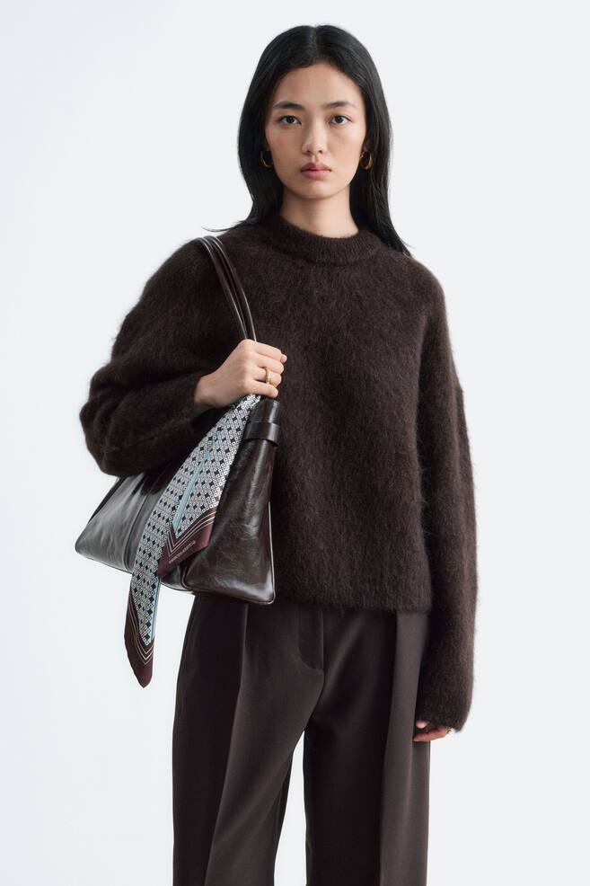 A dark brown mohair-blend jumper with a fuzzy, textured finish and a crew neck is being worn by the model. The jumper is styled with matching dark brown trousers, and the model is carrying a dark brown leather shoulder bag. - 1