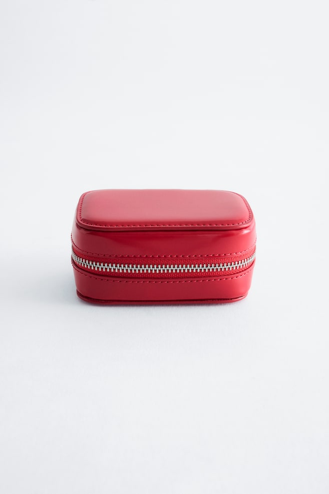 A small, rectangular red case with rounded corners is sitting facing forwards, featuring a smooth, semi-gloss finish with matching red topstitching. A silver-toned metal zip is wrapping horizontally around the centre of the structure. - 1