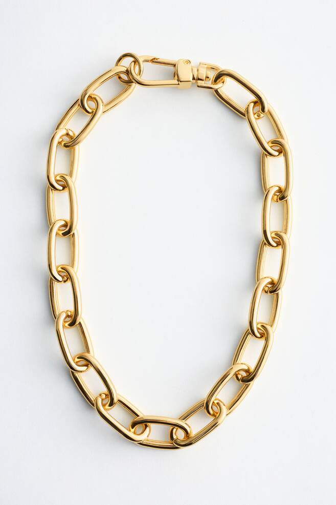 Het product is een goudkleurige, grove ketting met schakels. Hij is voorzien van grote, langwerpige ovale schakels en een karabijnsluiting die zichtbaar is en de uiteinden van de ketting met elkaar verbindt. - 1