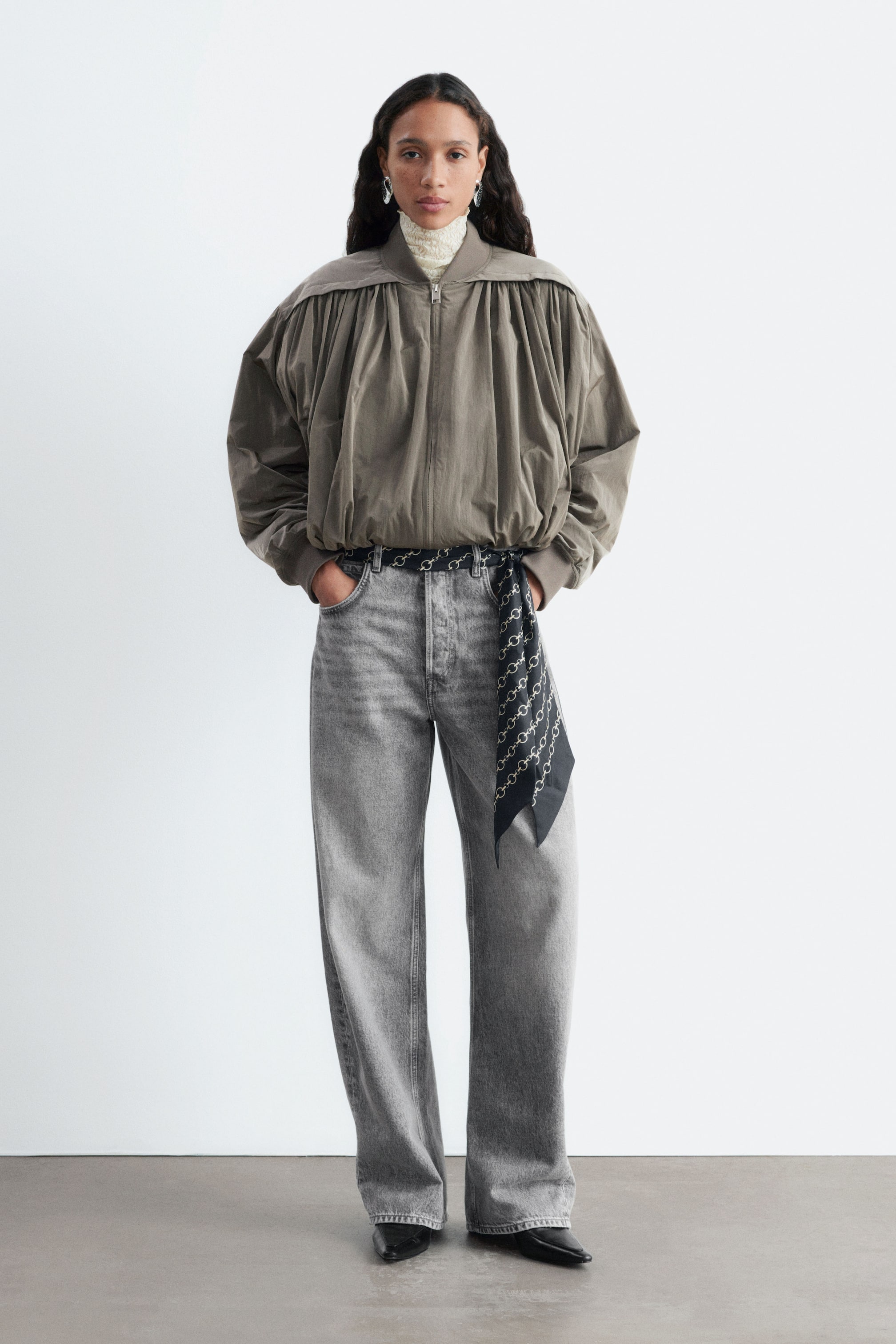 Agrandir l'image: Une veste bomber ballon taupe, présentant une silhouette volumineuse et arrondie avec des détails de tissu froncé sur la poitrine et les épaules, est portée par le mannequin. La veste zippée est associée à un jean gris taille haute et superposée sur un haut crème à col montant.