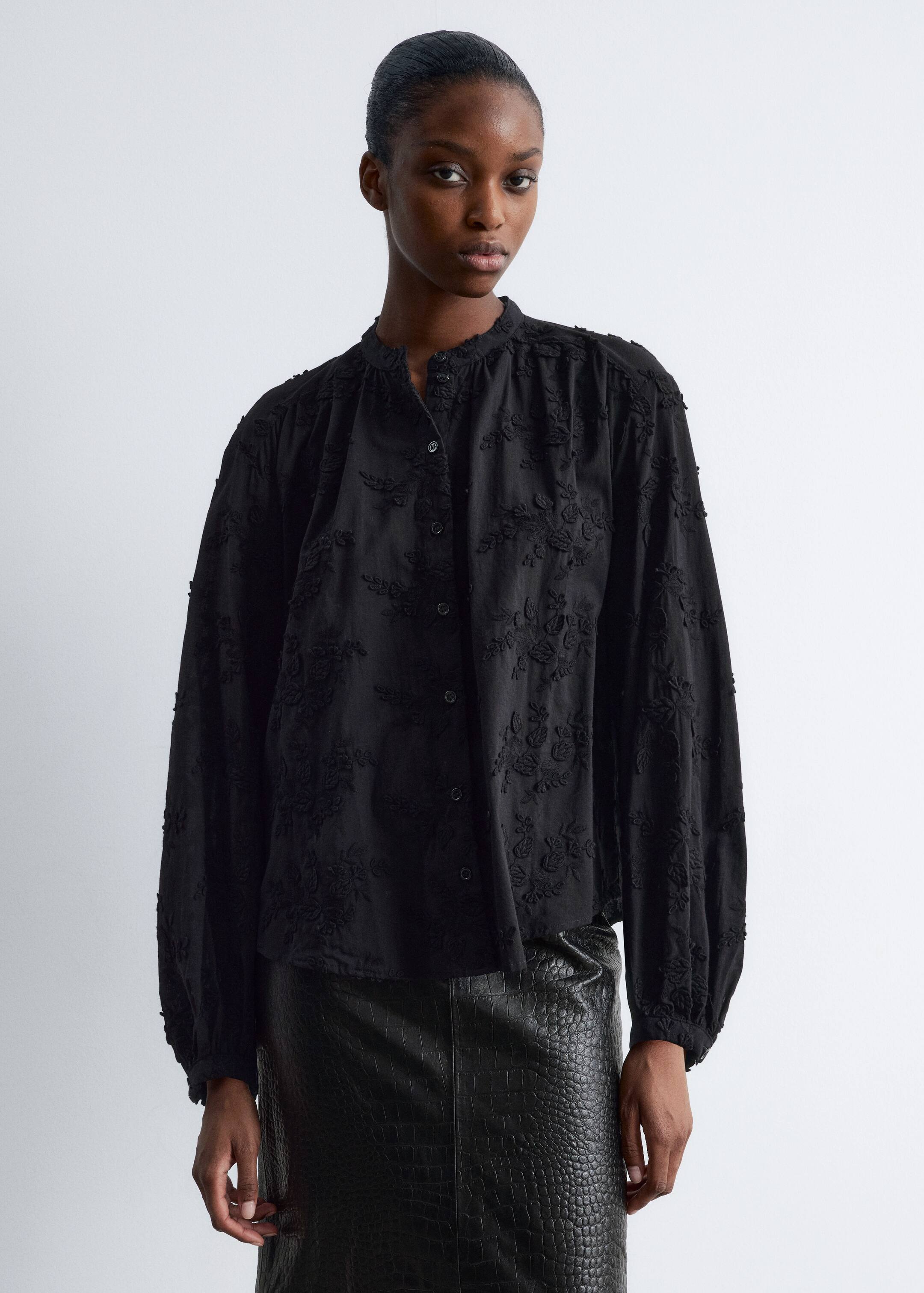 Ver imagem maior: Uma blusa preta de manga comprida está a ser mostrada de frente, apresentando um bordado floral texturizado em toda a peça. É desenhada com uma gola alta com folhos, uma carcela de botões à frente e mangas volumosas reunidas nos punhos, usada por fora de uma saia escura.