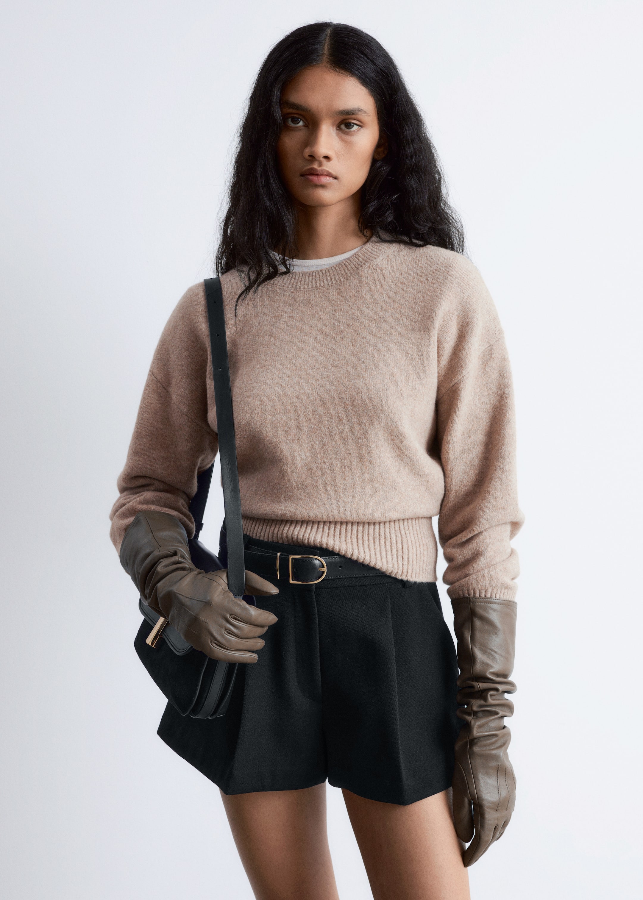 Größeres Bild anzeigen: Ein nach vorne gerichtetes Model trägt einen hellbraunen Pullover mit Rundhalsausschnitt, gerippten Bündchen und Saum, über einem weißen T-Shirt. Sie trägt auch hoch taillierte schwarze Shorts mit vorderen Falten, die von einem dünnen schwarzen Gürtel mit einer goldfarbenen rechteckigen Schnalle zusammengehalten werden. Der Look wird mit langen, dunkelbraunen Lederhandschuhen, die über ihre Ellbogen reichen, und einer kleinen schwarzen Schultertasche mit einem goldfarbenen Verschluss ergänzt.