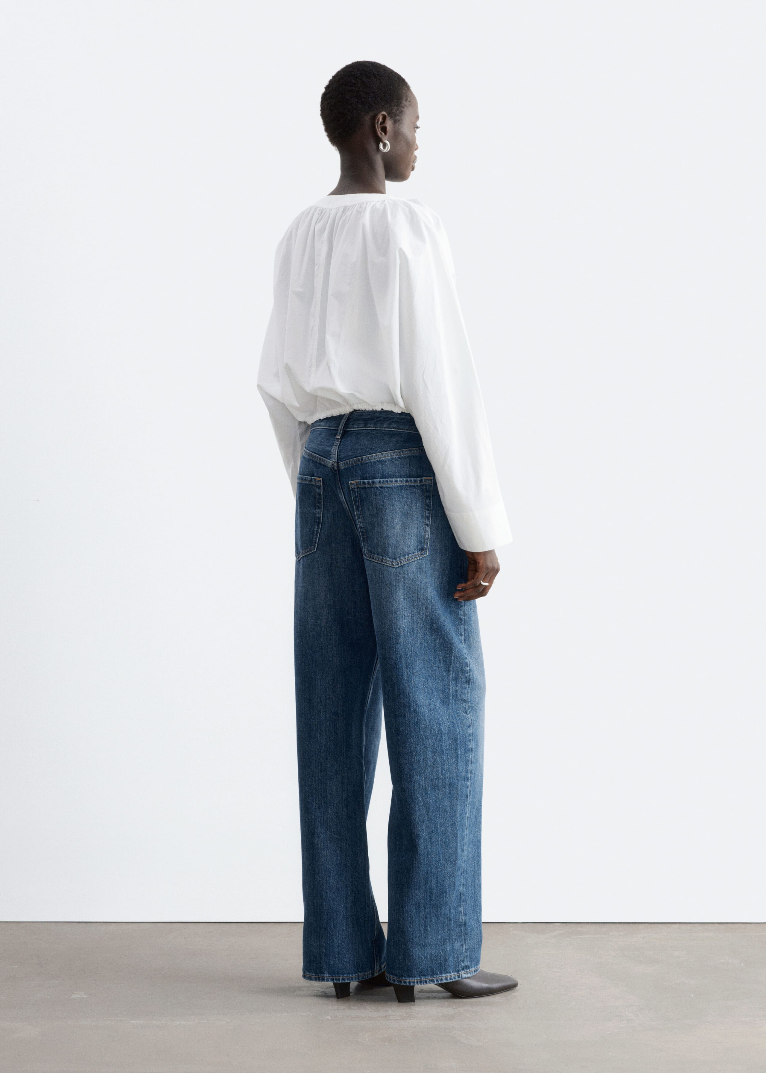 A white cropped cotton blouse featuring a gathered neckline and voluminous long sleeves is shown on the model from the back. The blouse is designed with a gathered drawstring hem and is styled with wide-leg blue jeans.