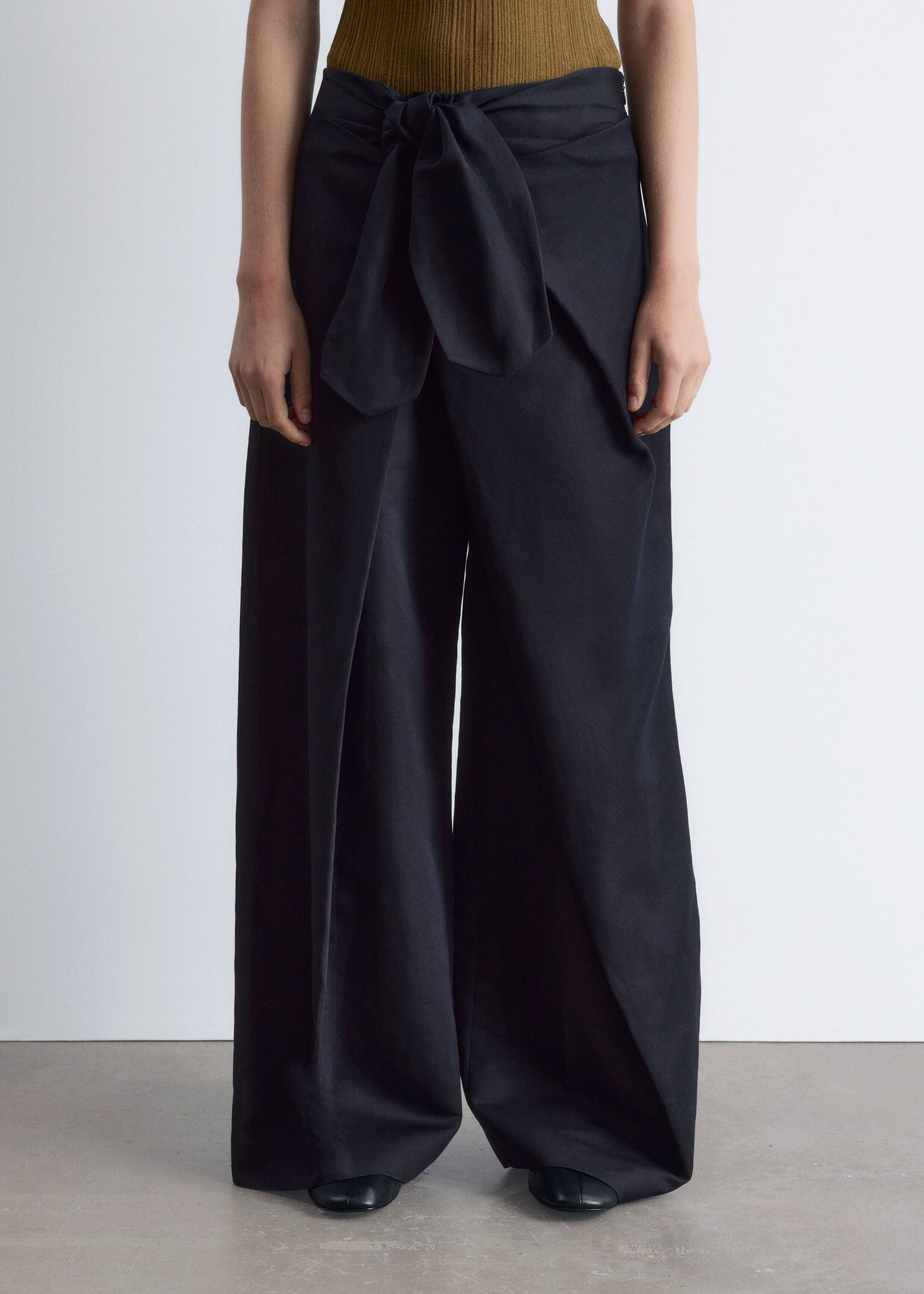Grotere afbeelding bekijken: Zwarte wijde broek wordt gepresenteerd vanuit een vooraanzicht, met een wijde, vloeiende snit en een opvallend strikdetail aan de voorkant van de taille. Ze worden gestyled met een geribd olijfgroen topje en zwarte flats.