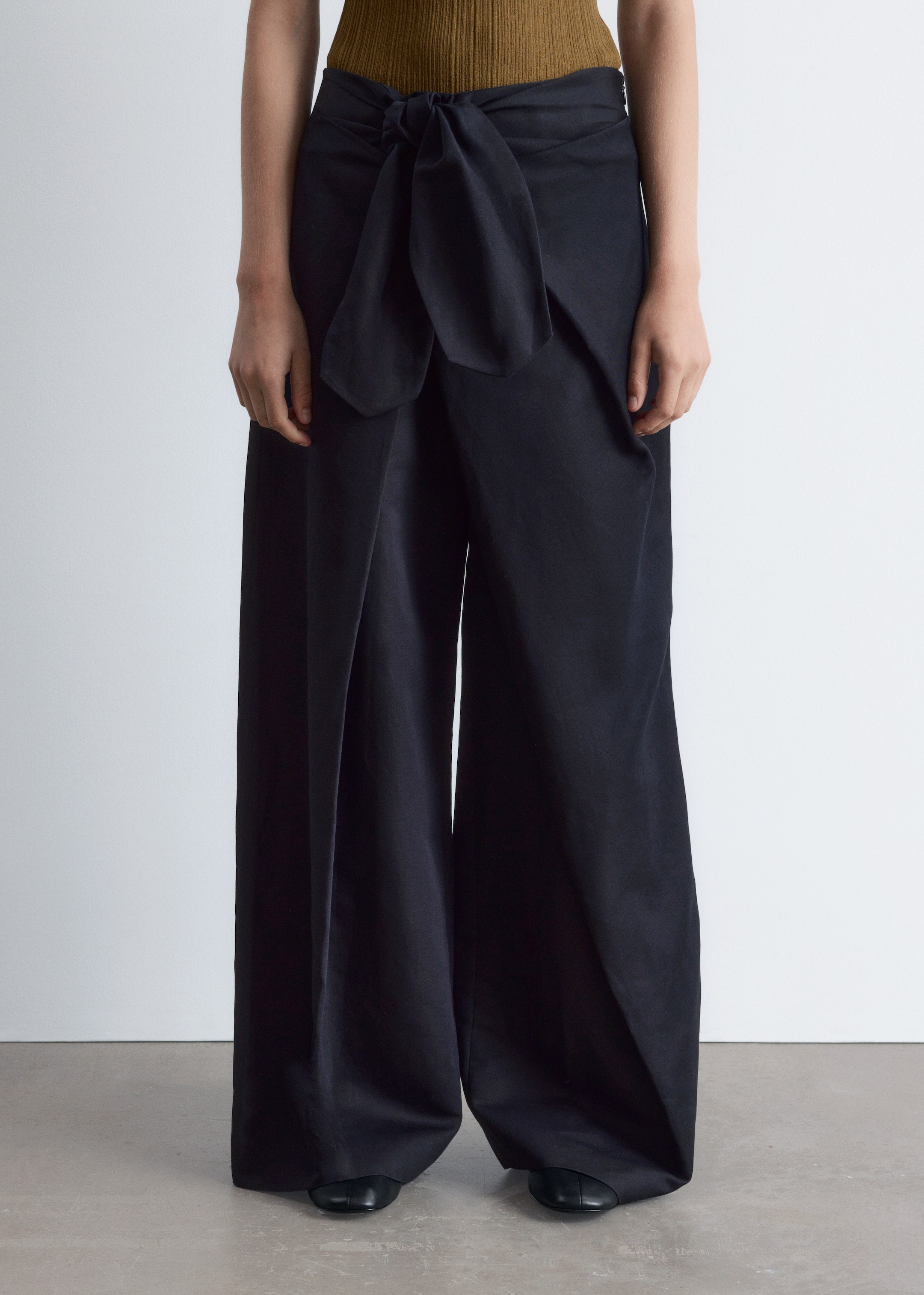 Agrandir l'image: Un pantalon noir à jambes larges est présenté de face, avec une coupe large et fluide et un détail de nœud papillon proéminent à la taille avant. Ils sont stylisés avec un haut côtelé vert olive et des ballerines noires.