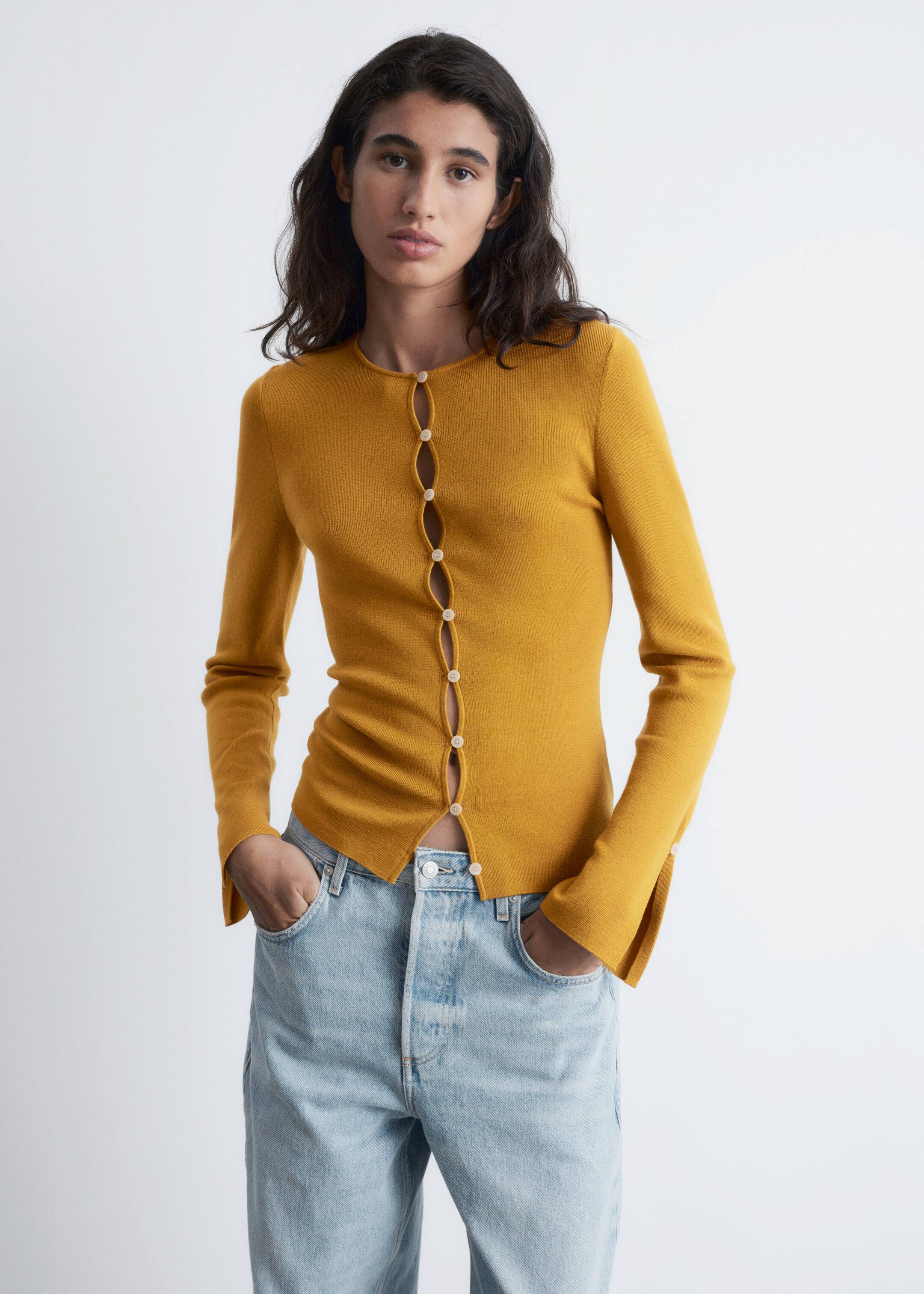 Agrandir l'image: Un haut en maille fine de couleur jaune moutarde est présenté, avec un col rond et une patte de boutonnage avant unique avec des boutons espacés de couleur claire qui créent un design ondulé et ouvert. Le haut ajusté à manches longues est partiellement déboutonné en bas et se termine par des poignets fendus subtils.
