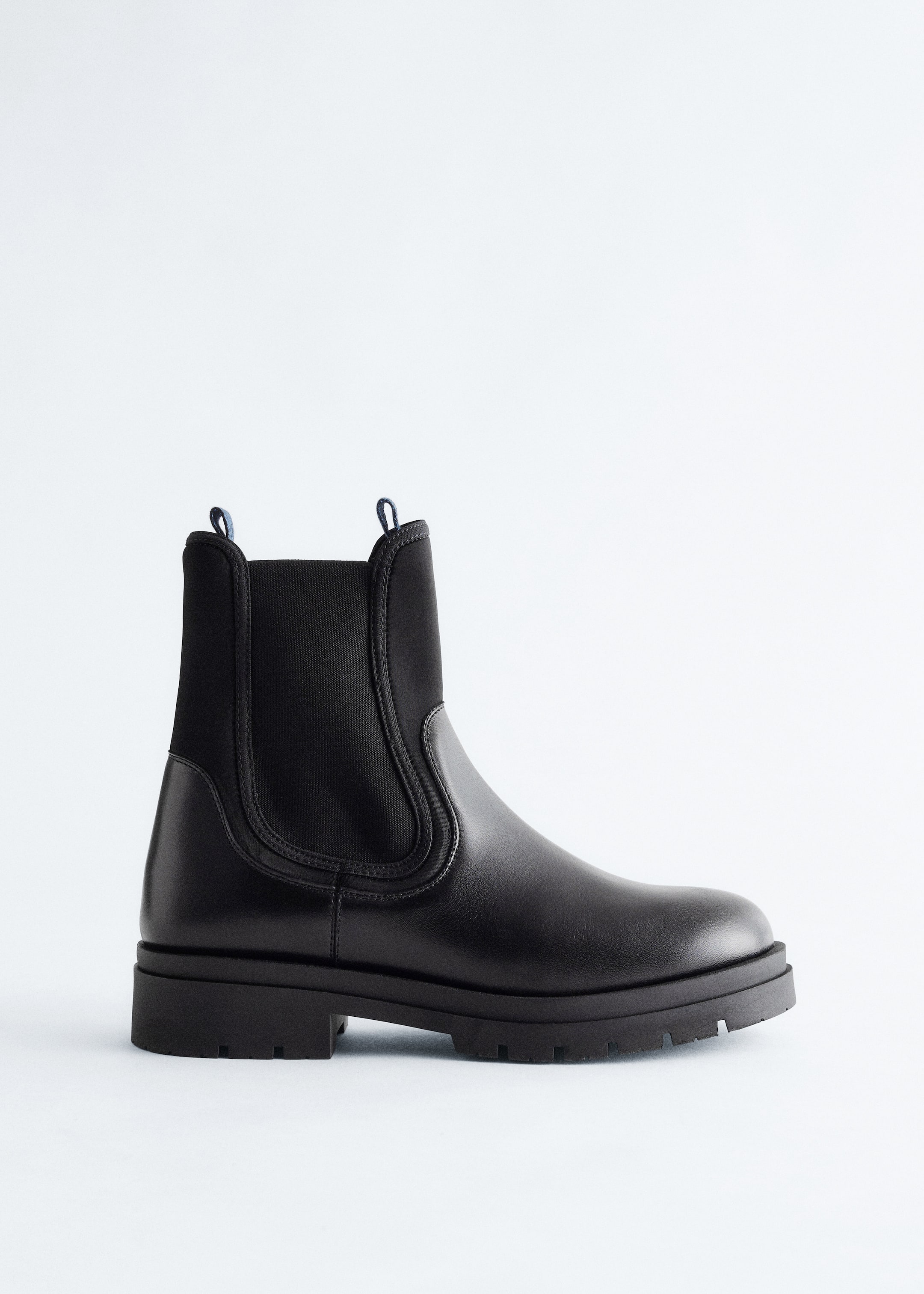 Ingrandisci l'immagine: Un Chelsea boot nero è presentato, rivolto a sinistra, con un piede in pelle liscia e un gambale in scuba con inserti elastici laterali. Due linguette di trazione azzurro chiaro sono visibili nella parte superiore del gambale, e lo stivale è posizionato su una suola spessa e modellata.