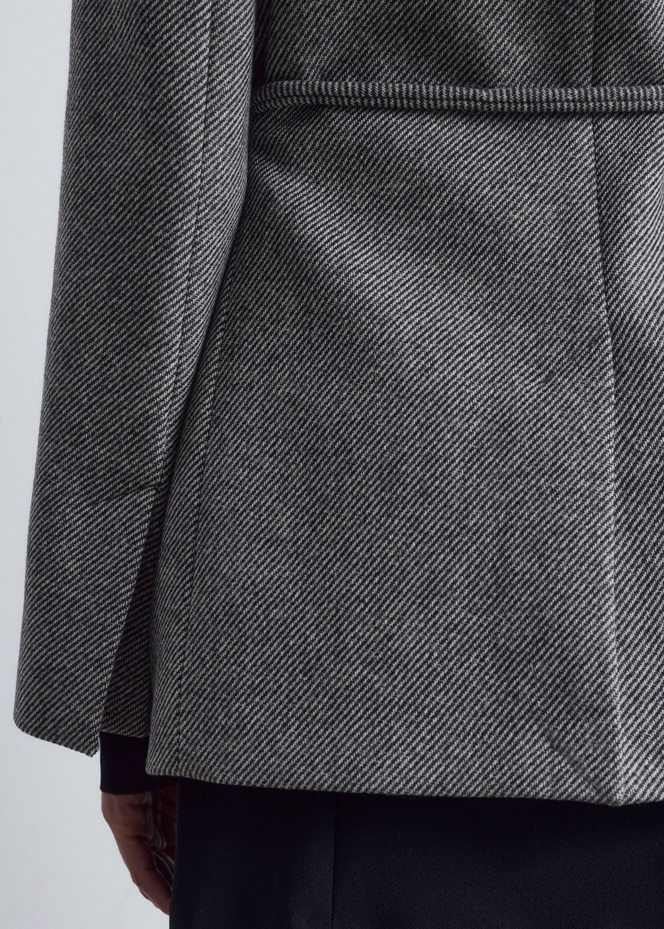 Agrandir l'image: Une veste en chevrons gris et noir est présentée de dos, mettant en valeur son design structuré. La veste présente des coutures verticales sur le panneau arrière et un détail de demi-ceinture horizontale, avec sa manche gauche affichant une couture proéminente. Un vêtement sombre est visible sous l'ourlet de la veste.